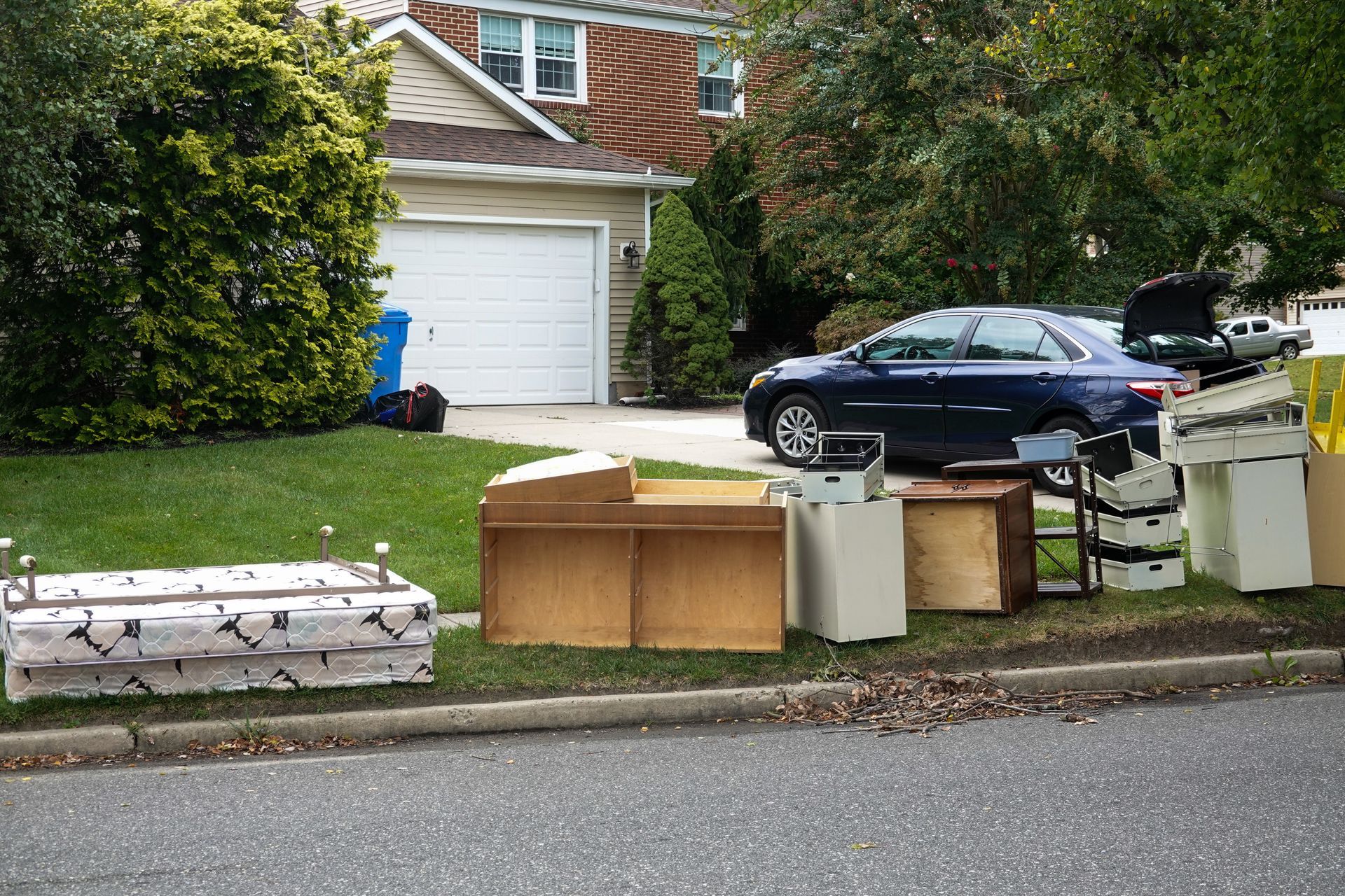 Cardboard boxes and a white appliance piled along a residential curb beside parked cars and a garage.