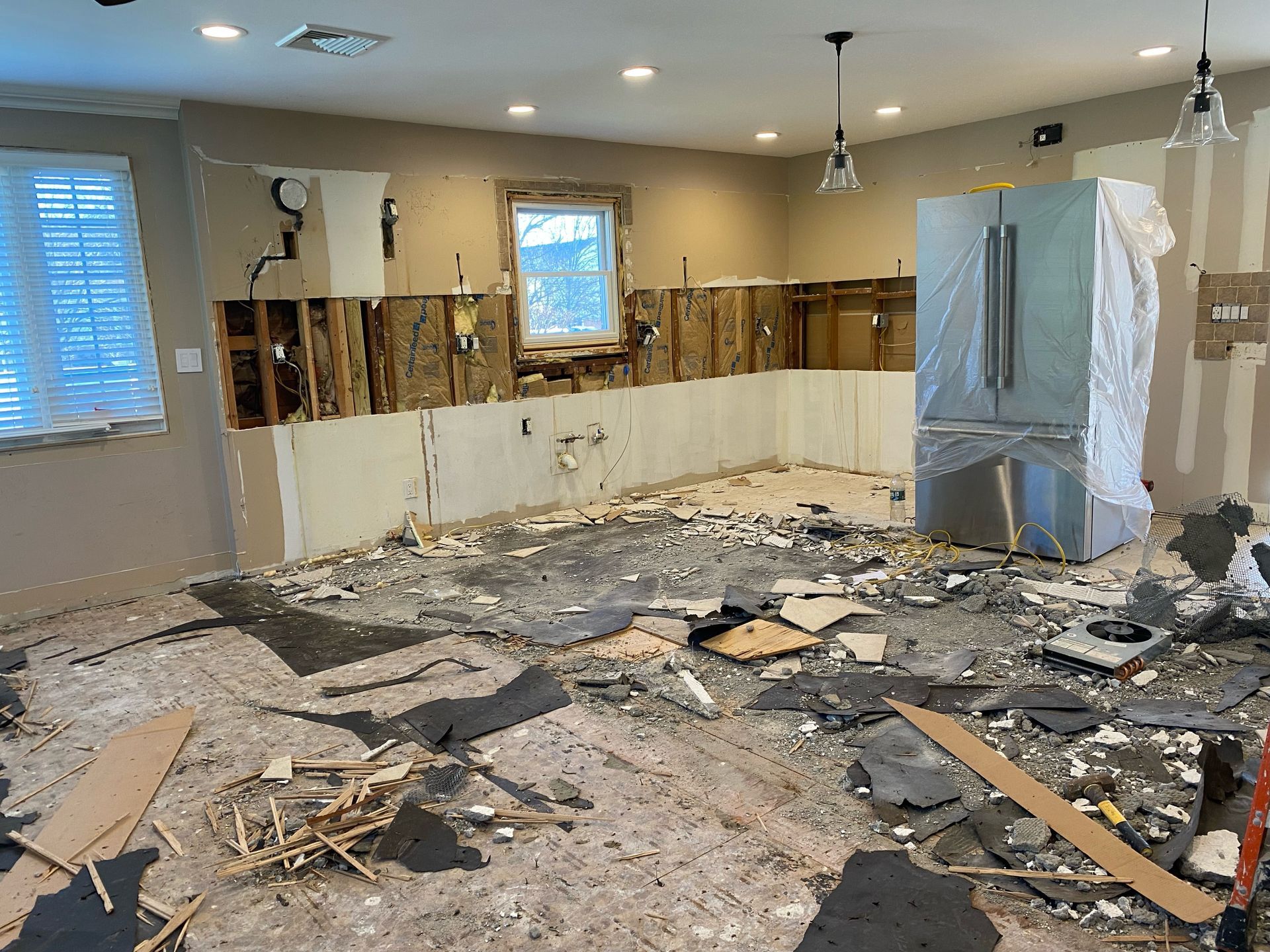 Flood-damaged kitchen with torn-out cabinets, debris on the floor, and a refrigerator standing amid renovations.