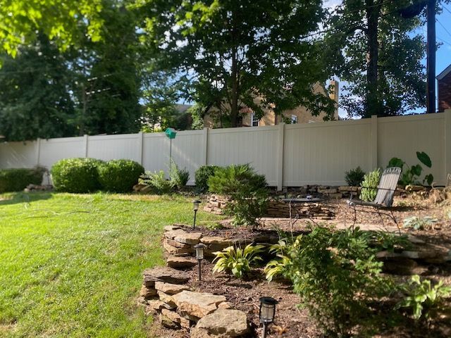 Backyard with white fence, stone landscaping, lawn, and greenery.