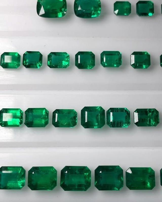 Rows of various-sized, rectangular and square, vibrant green emerald gemstones arranged on a white surface.
