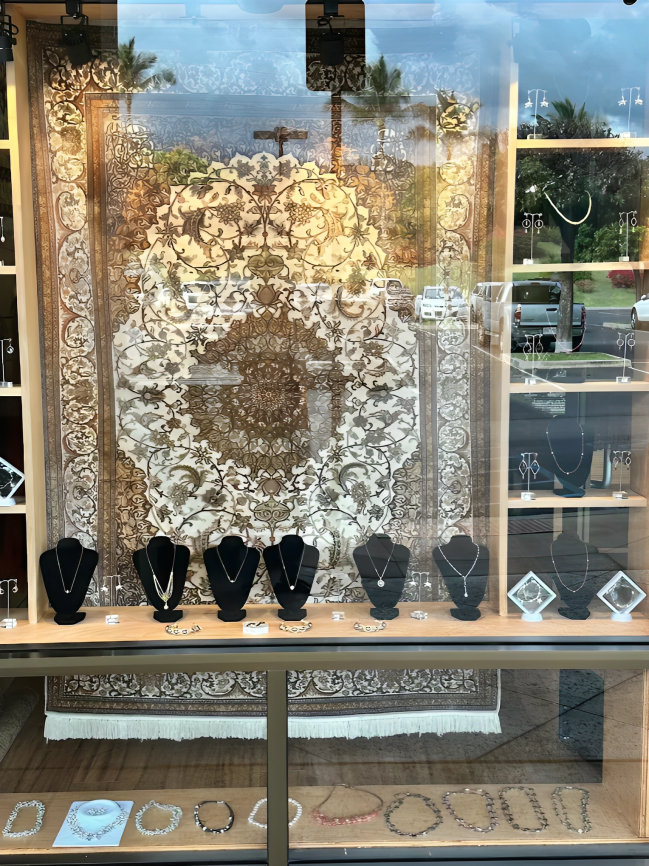 Jewelry store window display featuring necklaces on black stands, with a patterned rug backdrop.