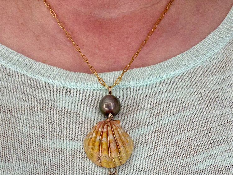 Woman wearing a gold necklace with a shell pendant and dark pearl, over a light blue sweater.