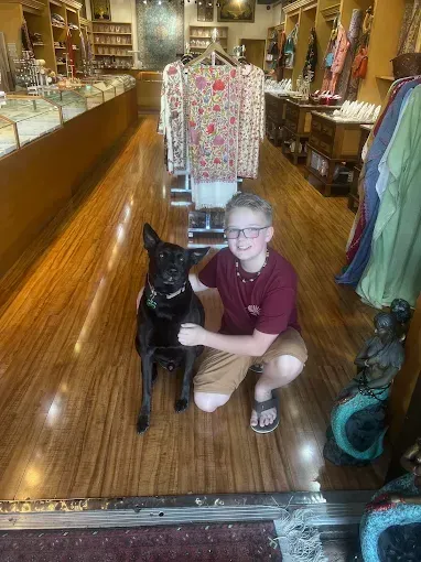 Boy kneels next to a black dog inside a shop with a wooden floor, smiling.