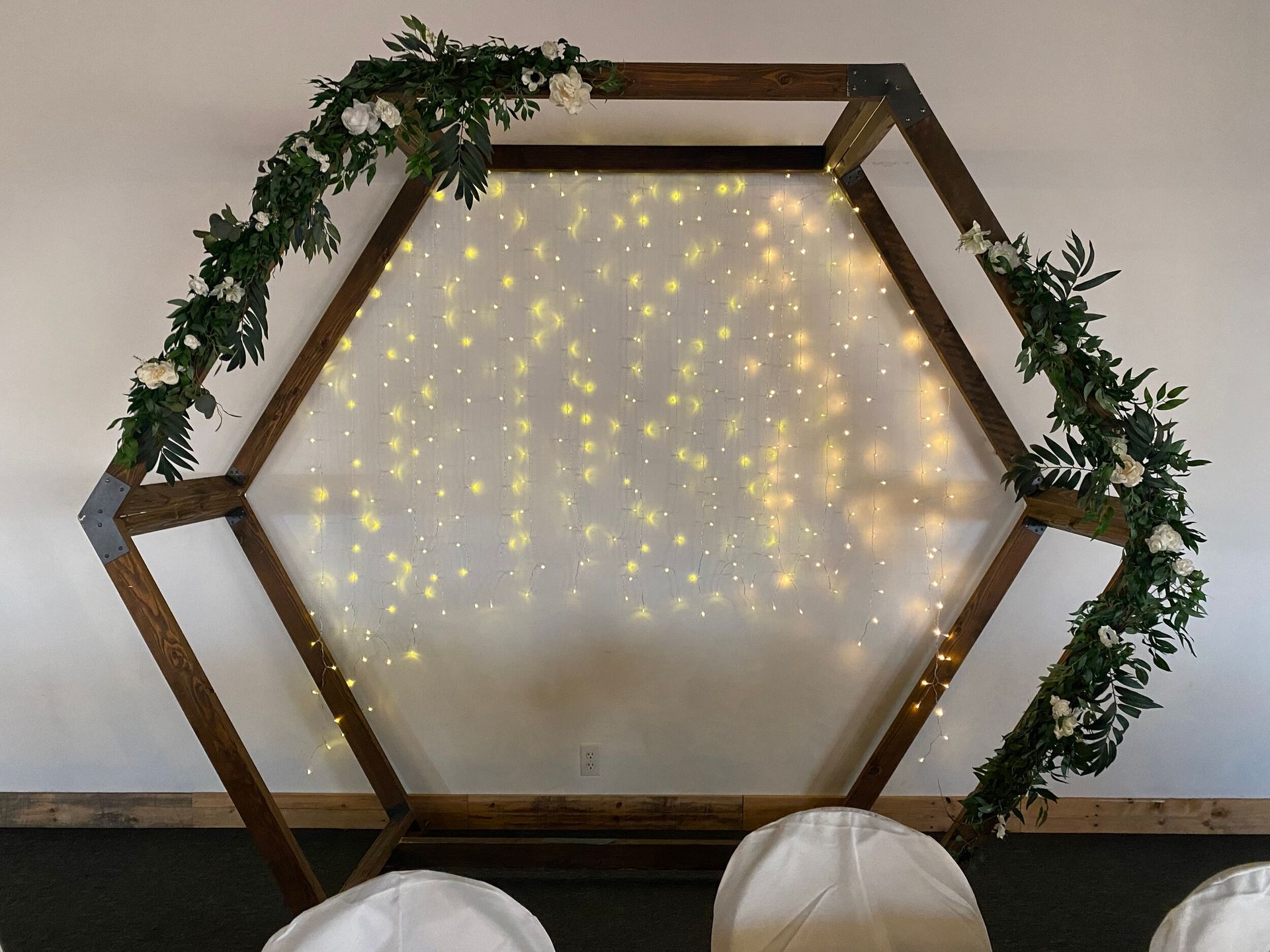 A wooden arch with flowers and lights on it is in a room with chairs.