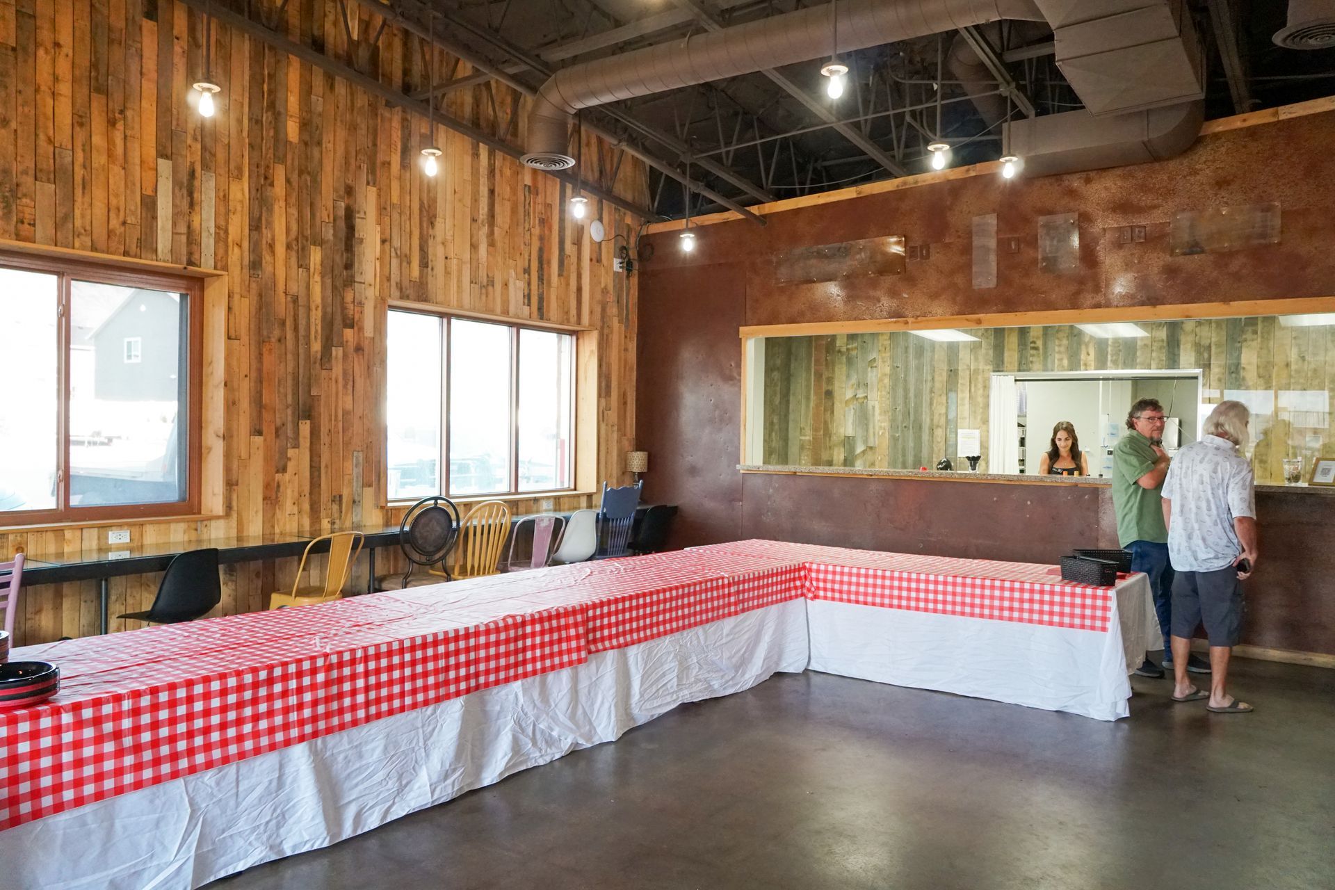 Interior of a building with a long table covered in red checkered cloth. People stand near a counter.