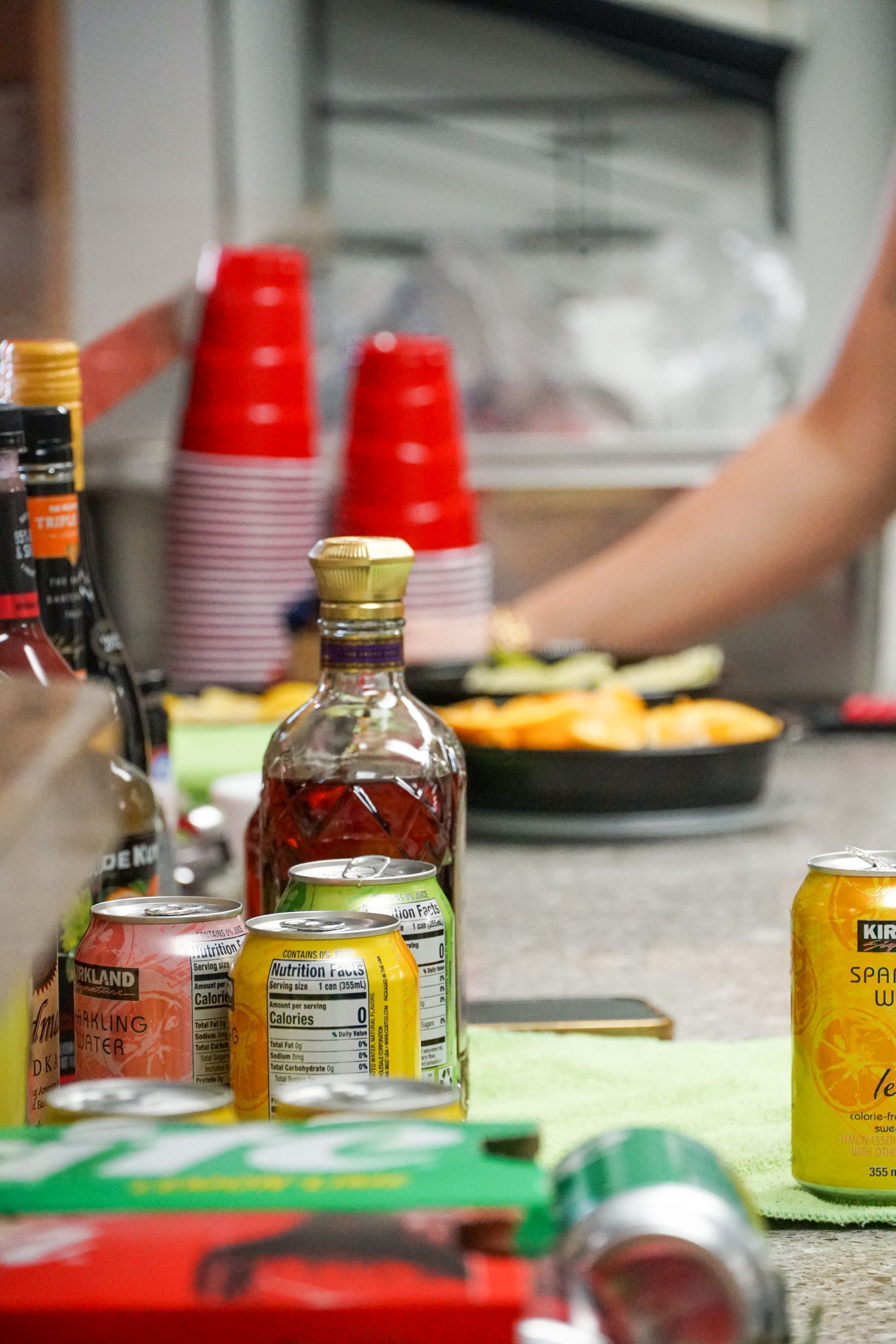 Bar setup with liquor bottles, canned drinks, red cups, and a tray of food on a counter.