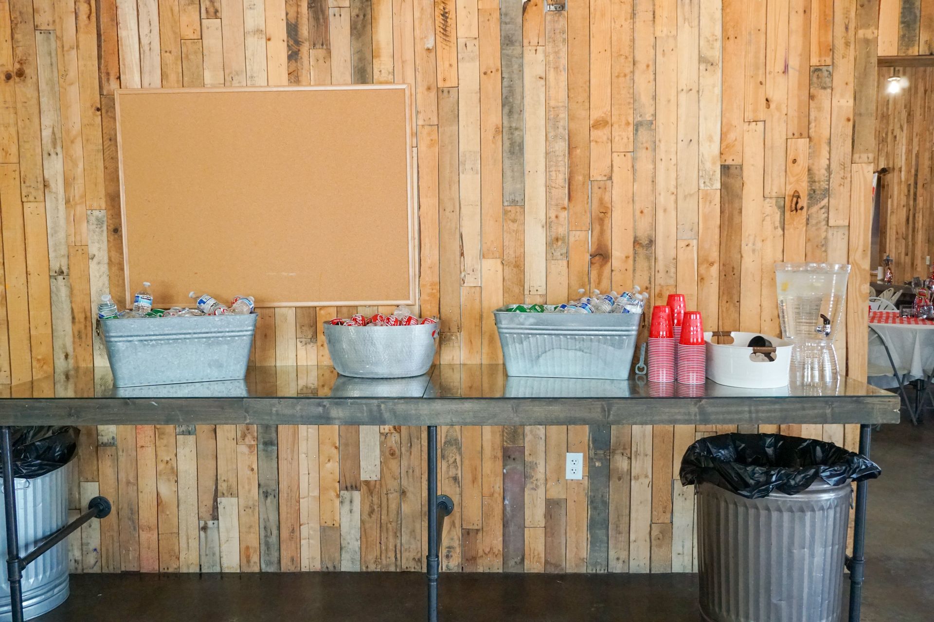 Beverage station with wooden wall backdrop, metal containers, drinks, cups, and trash cans.