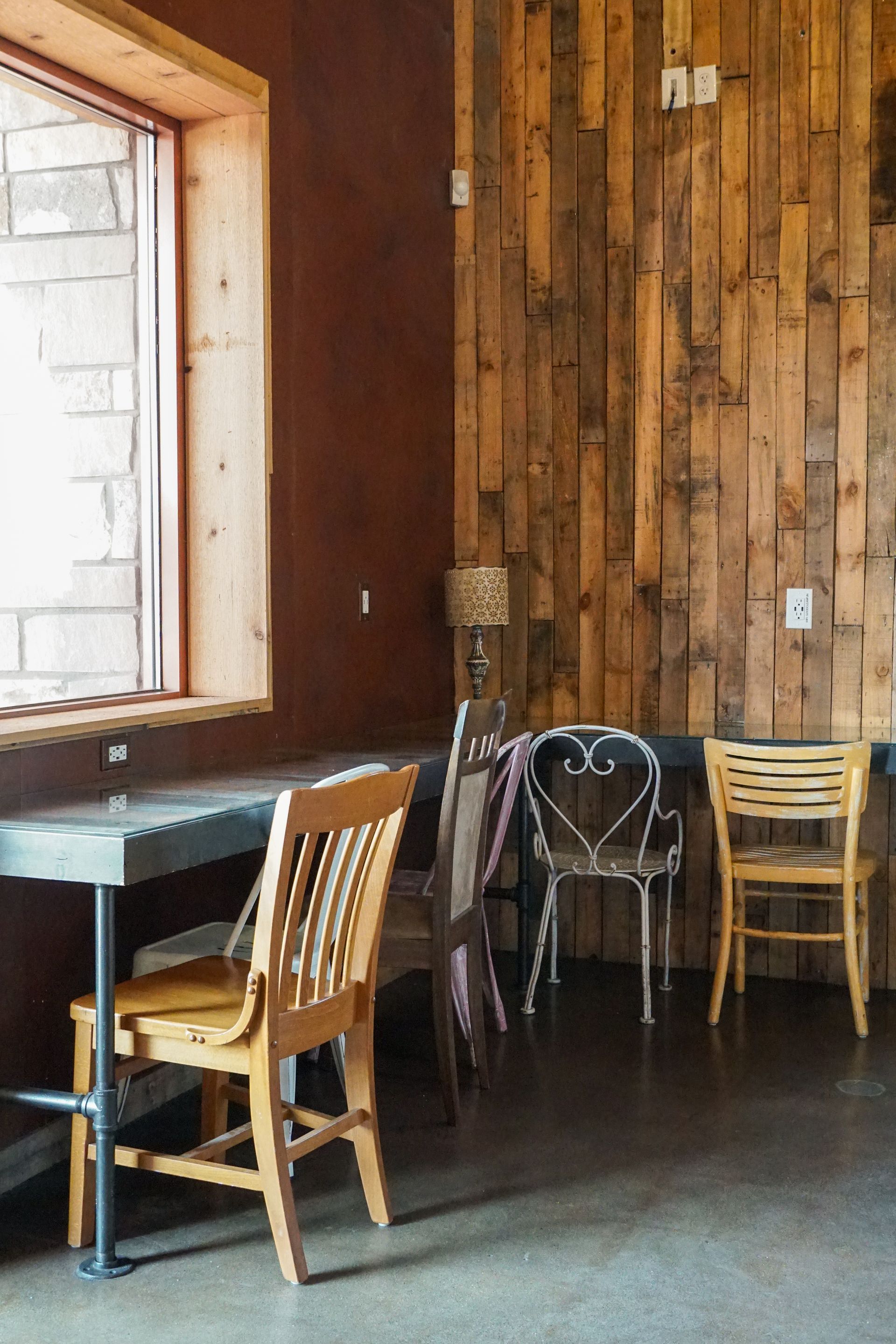 A cafe interior with tables, chairs, a wooden wall, and a window. Two people.