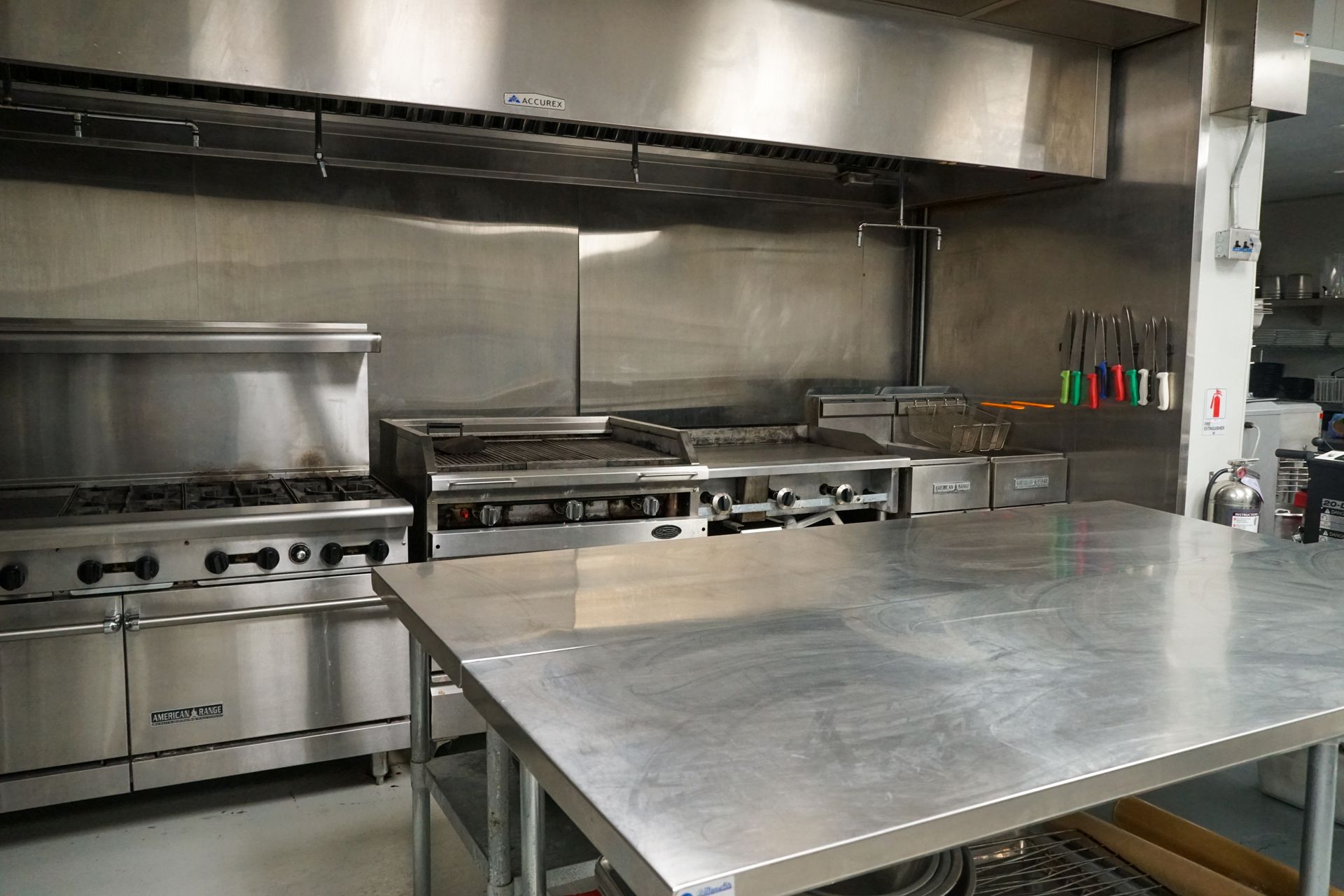 Stainless steel commercial kitchen with stove, grill, table, and vent hood.