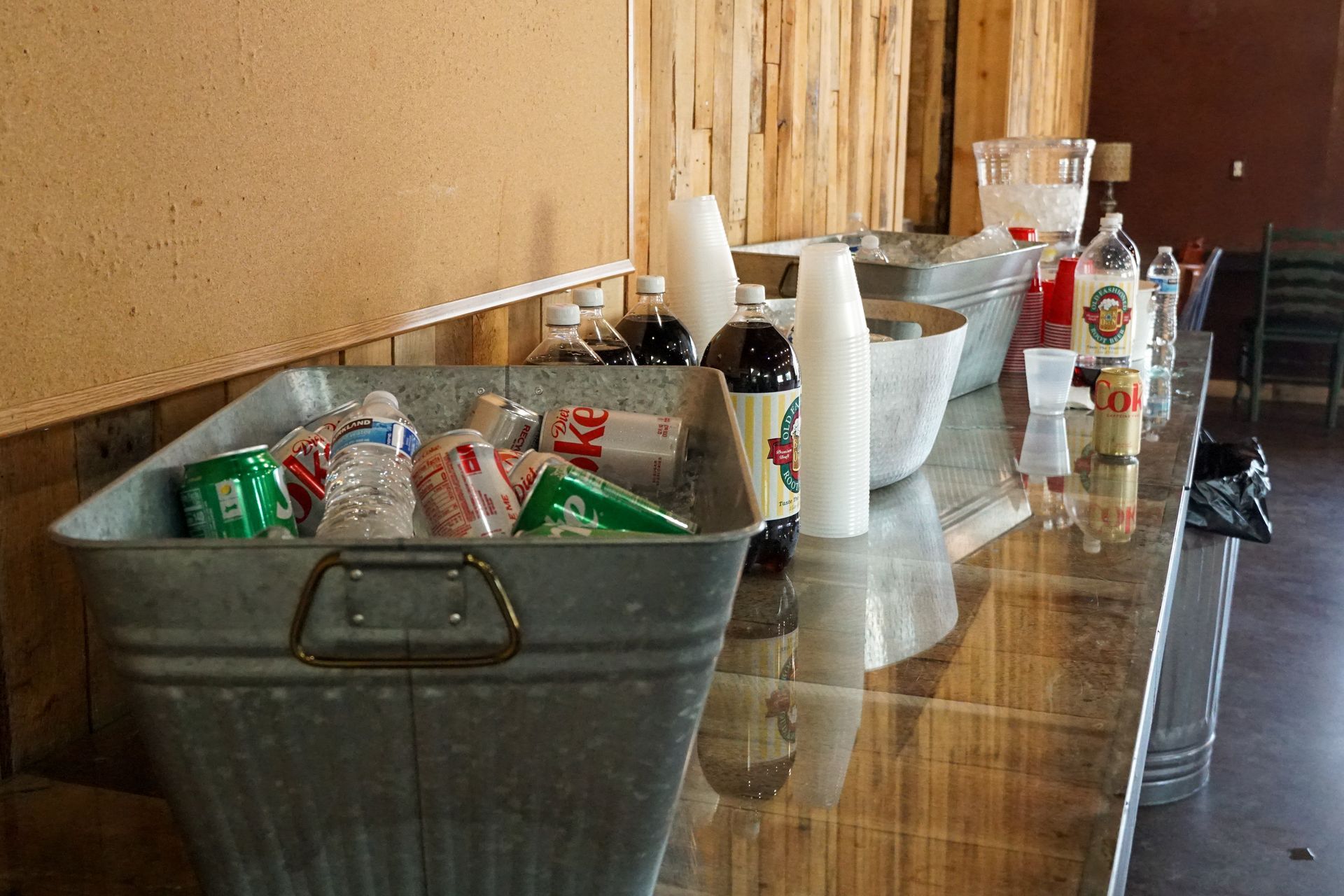 Metal tub filled with drinks on a counter, bottles and cups nearby. Rustic wood paneling in background.