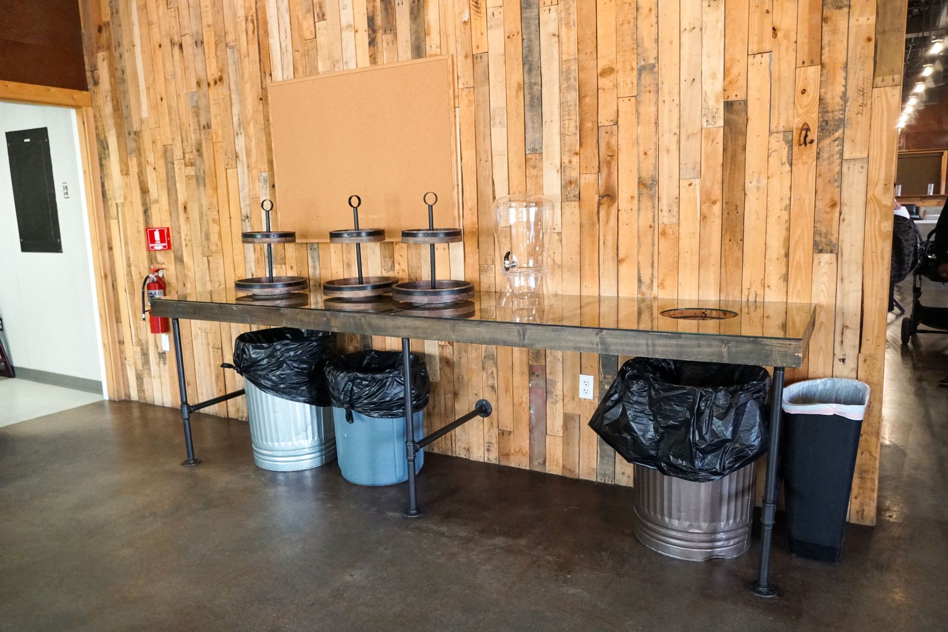 Long wooden table with three beverage dispensers and trash cans against a wood-plank wall.