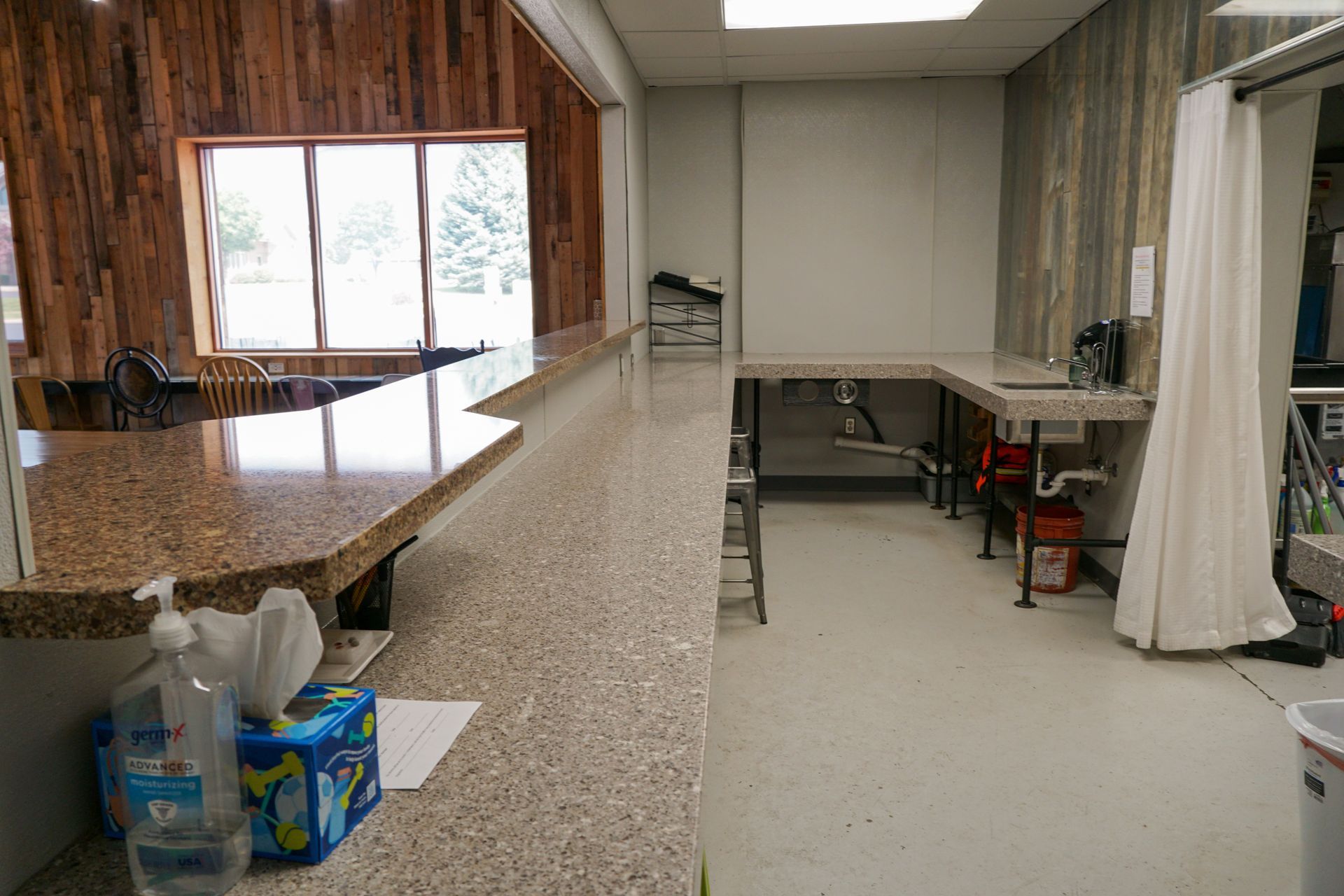 Room with a countertop, window, and a wash station. The decor includes wood paneling and a white curtain.