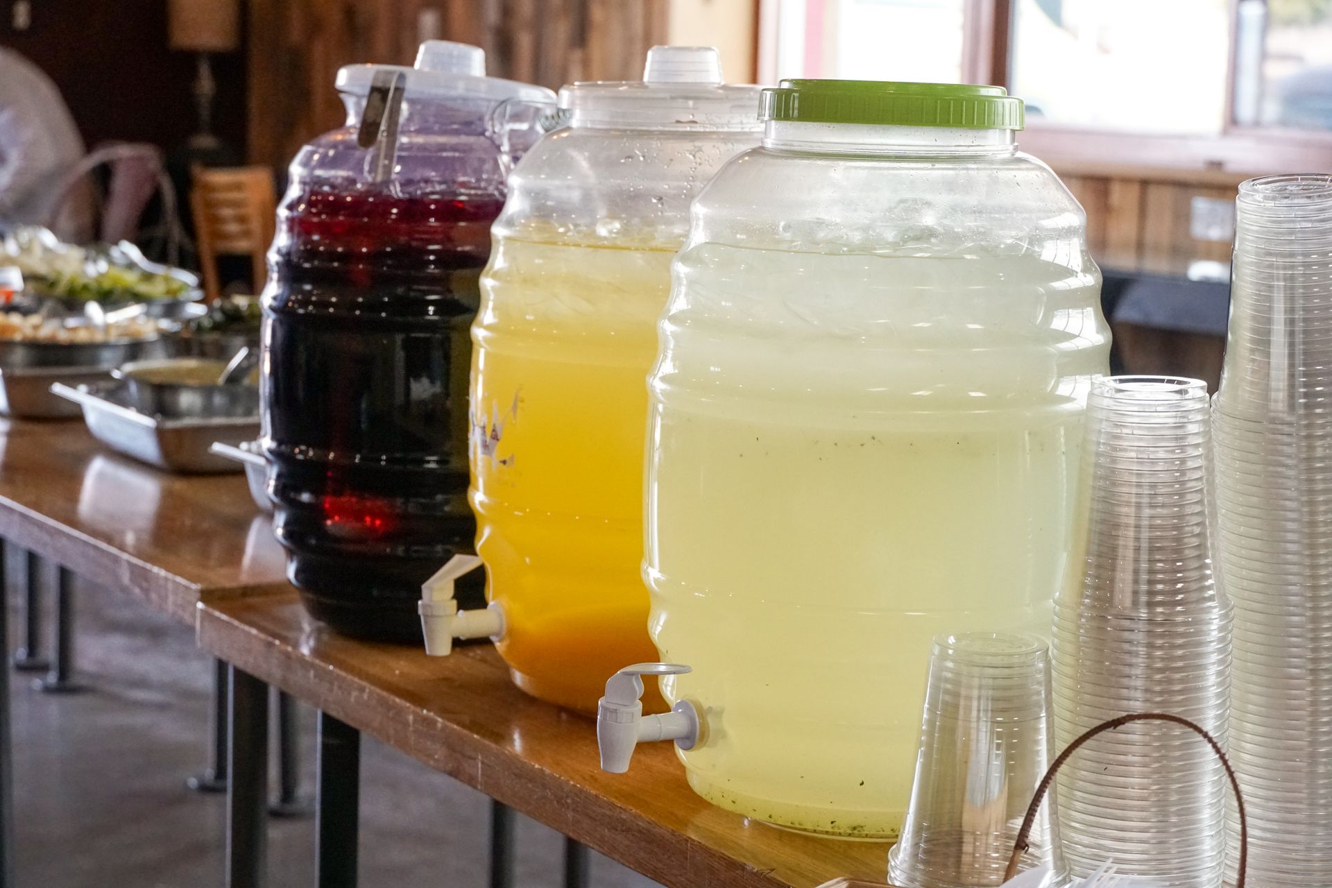 Three beverage dispensers with purple, orange, and yellow drinks, next to a stack of cups.