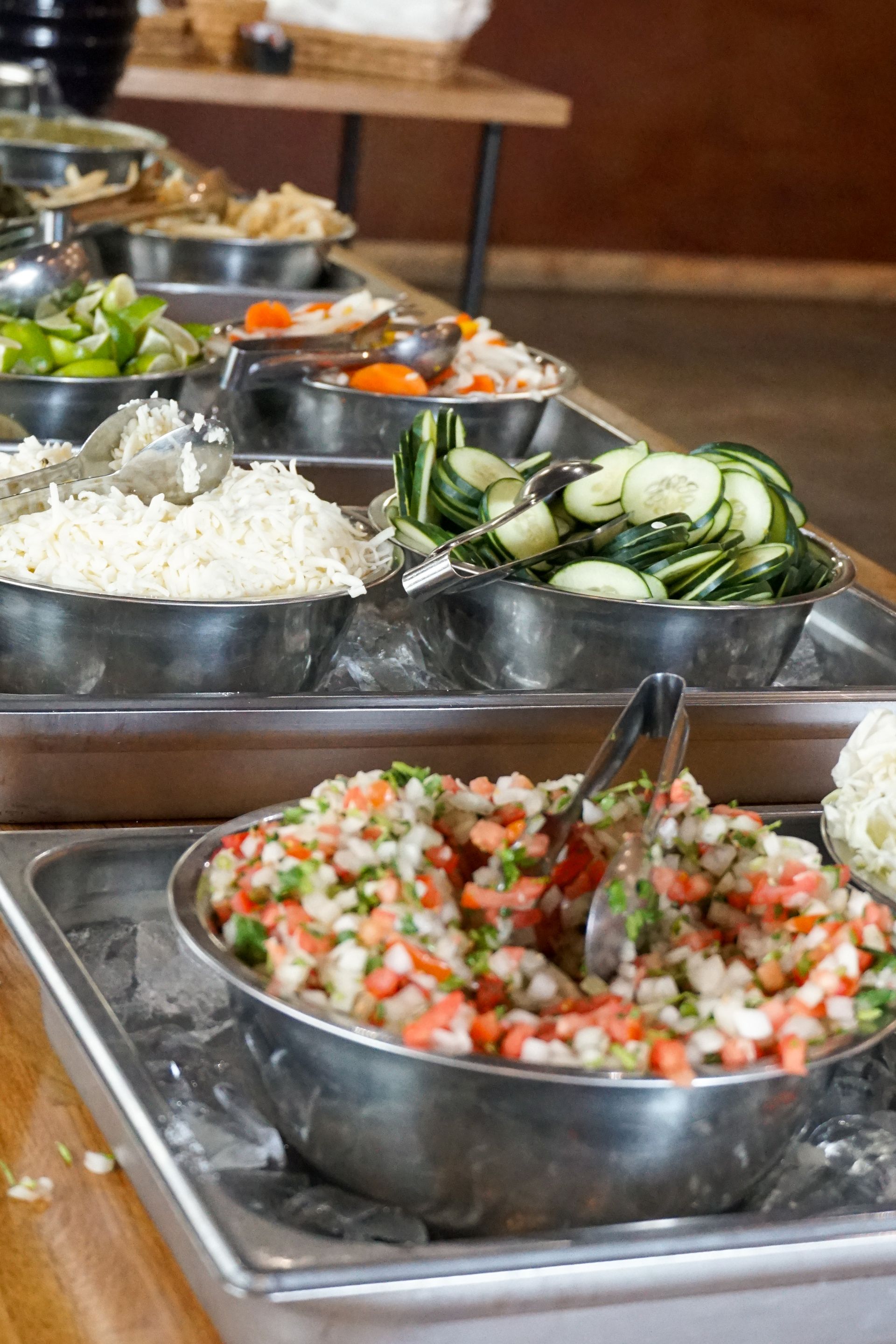 Salad bar with various toppings in metal containers, including chopped vegetables and cucumbers.