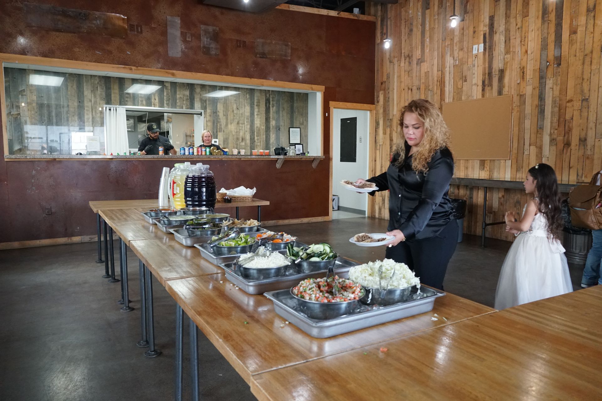 A woman serving food from a buffet in a rustic hall; other people in the background.