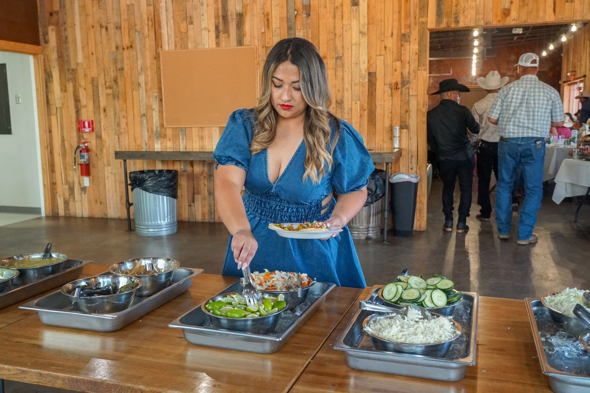 Woman in blue dress serves food at a buffet table in a rustic venue; other people in the background.
