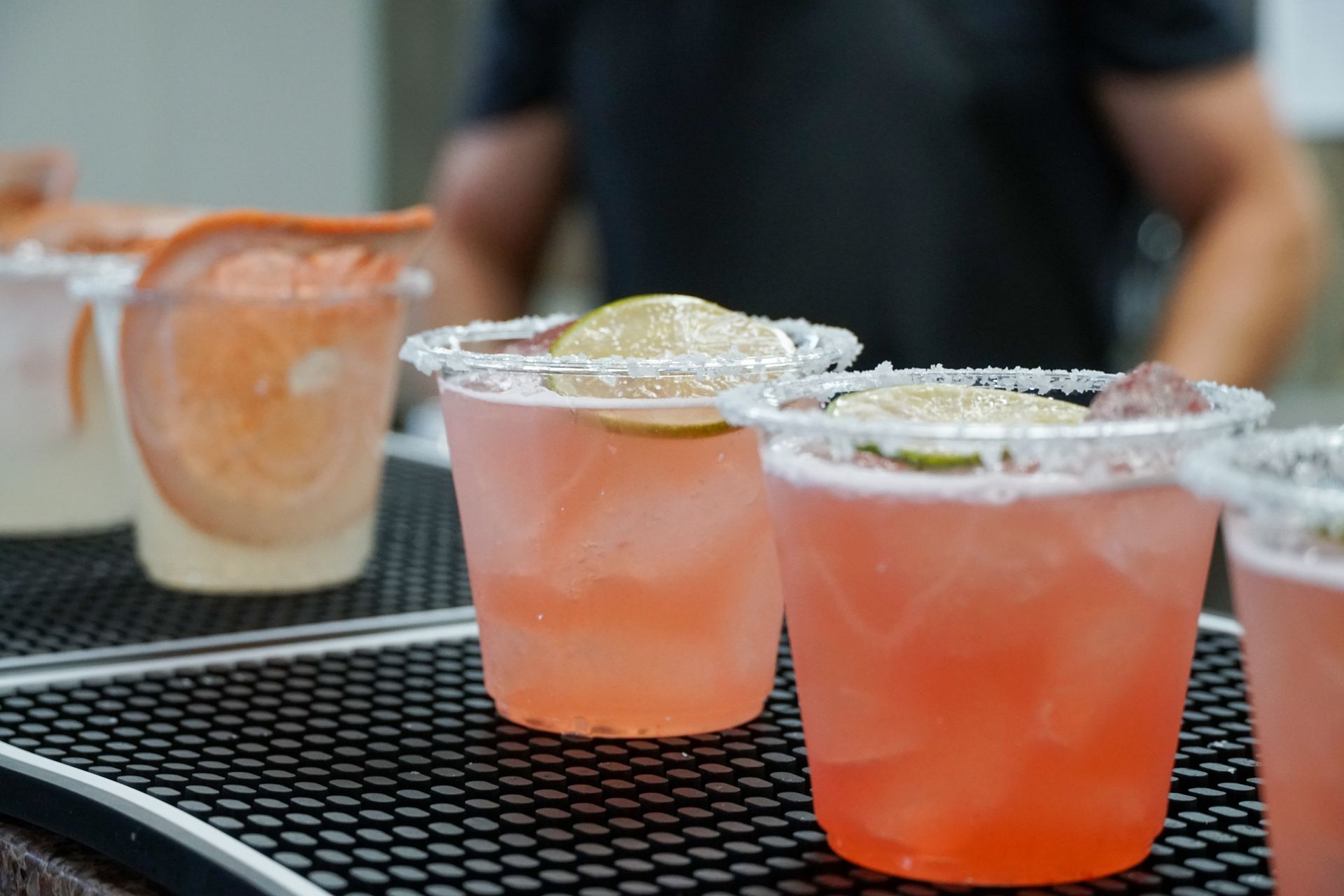 Close-up of four pink cocktails in plastic glasses with salt rims and lime slices, on a black bar.
