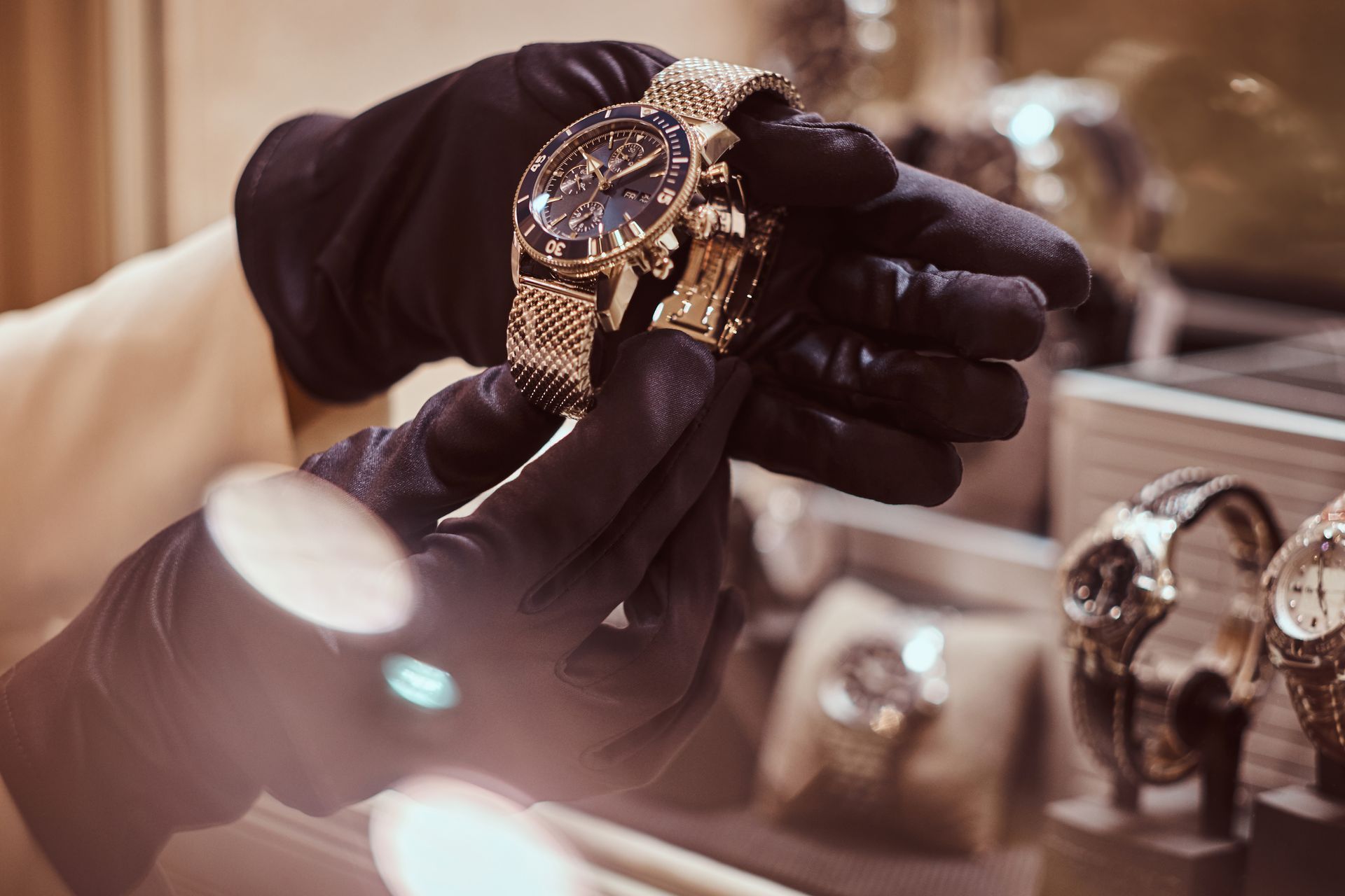 Person wearing black gloves holding a gold wristwatch with a dark blue face in a display case.