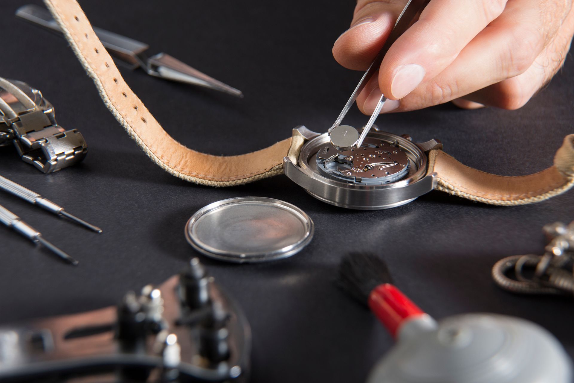 Person changing a watch battery with tweezers, surrounded by watch repair tools on a black surface.