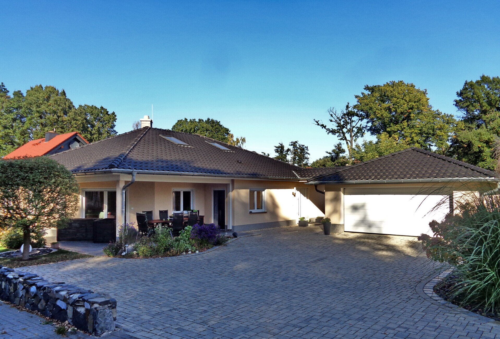 Beigefarbenes, eingeschossiges Haus mit schwarzem Dach, angebauter Garage und gepflasterter Auffahrt. Umgeben von Bäumen und Pflanzen.