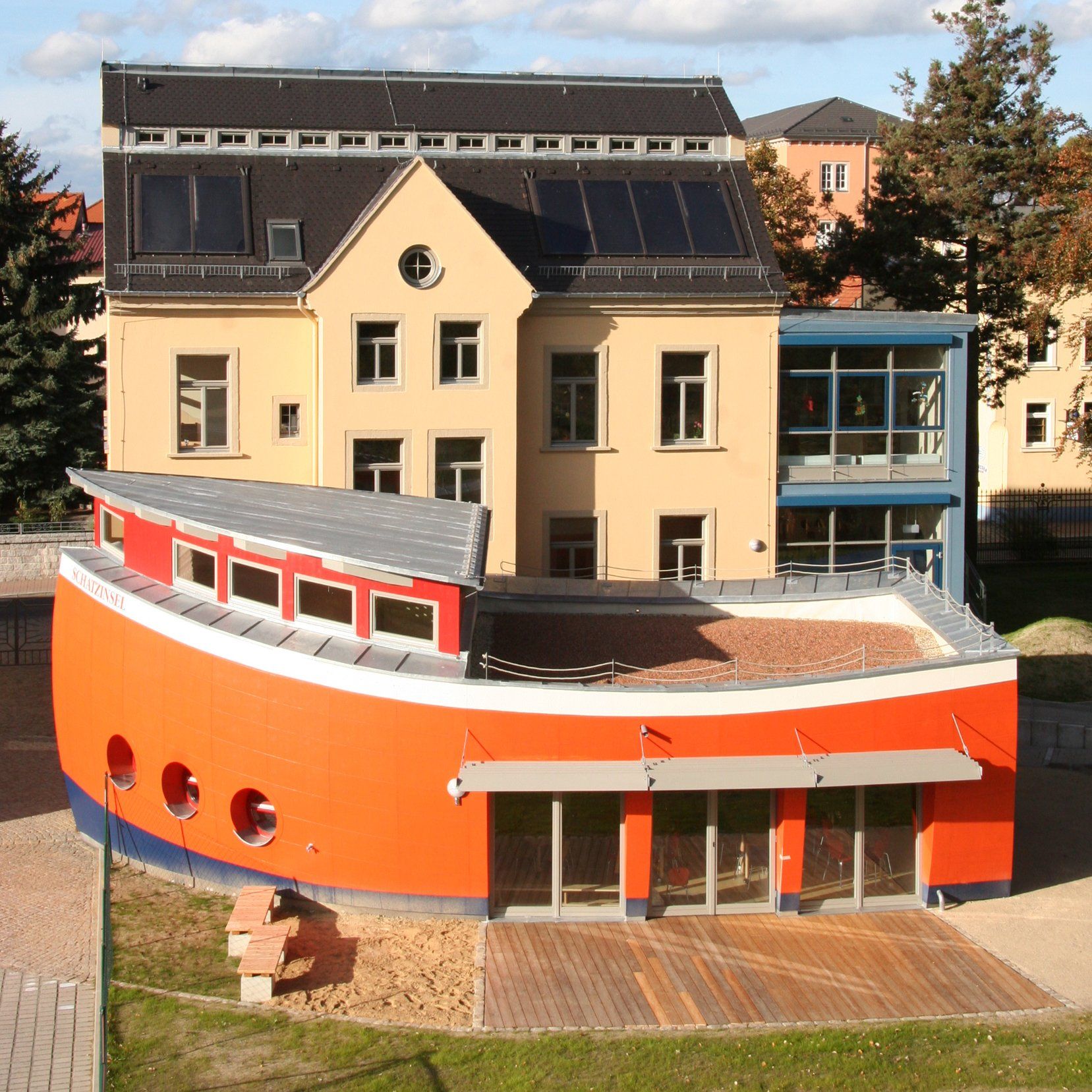 Ein orangefarbenes, bootsförmiges Gebäude mit runden Fenstern steht vor einem hellbraunen Gebäude mit Solarpaneelen auf dem Dach.