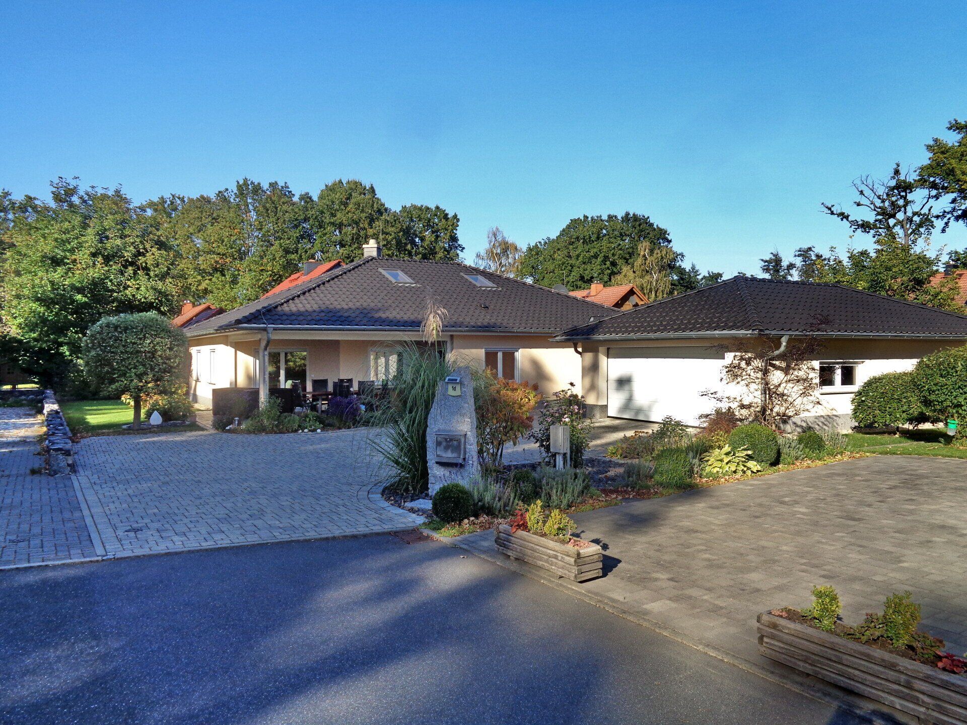 Ansicht aus der Froschperspektive auf ein beigefarbenes, eingeschossiges Haus mit schwarzem Dach, Steinauffahrt und umgeben von Bäumen unter klarem, blauem Himmel.
