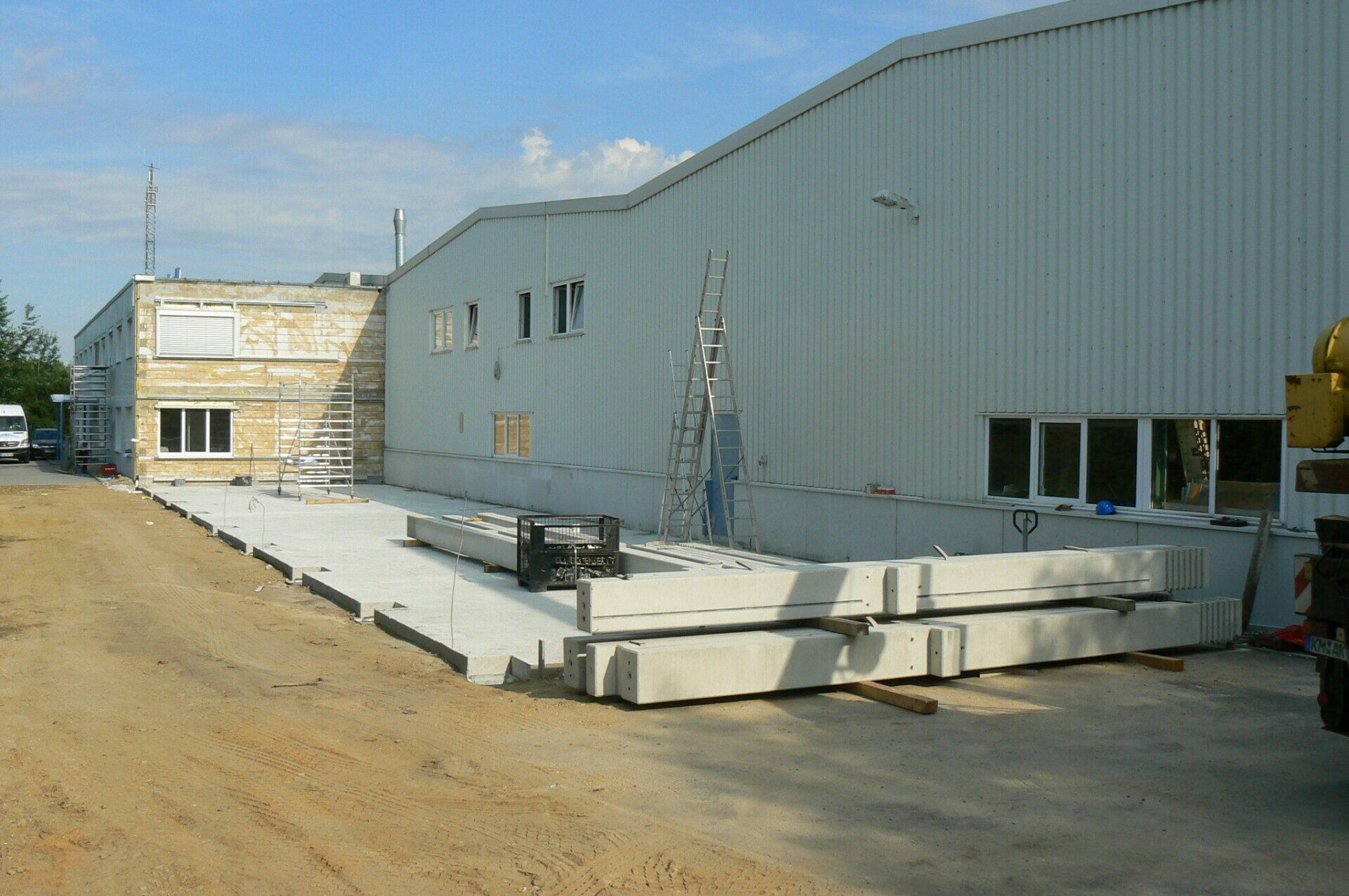 Außenansicht eines im Bau befindlichen Gebäudes mit Betonplatten, Wellblechverkleidung und umgebender Erde.