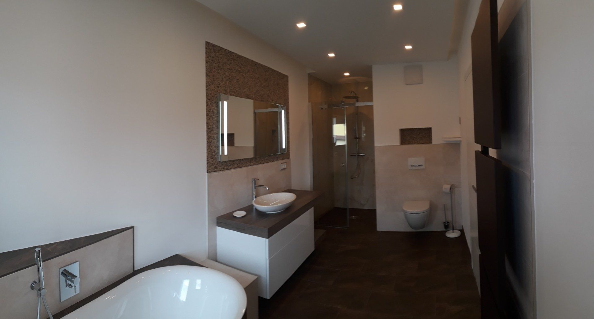 Modernes Badezimmer mit weißem Waschbecken, WC und Badewanne. Beige Fliesen und dunkler Holzboden.