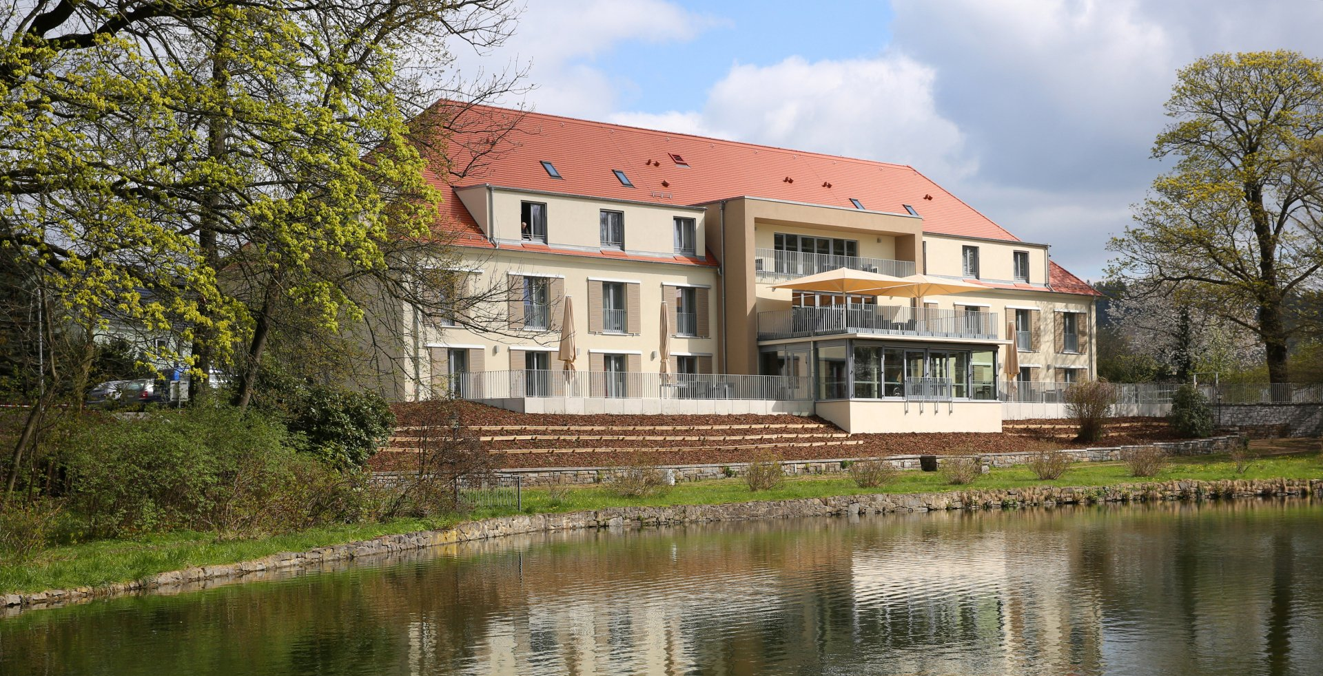 Großes Gebäude mit rotem Ziegeldach an einem See. Terrassenförmiger Bereich. Sonniger Tag.