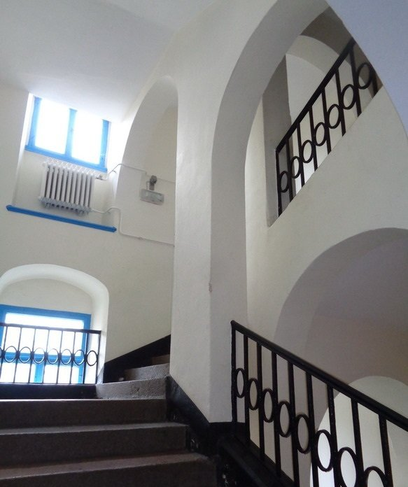 Treppenhaus mit weißen Wänden, Bogenöffnungen und schwarzem schmiedeeisernem Geländer. Fenster mit blauem Rahmen sind sichtbar.