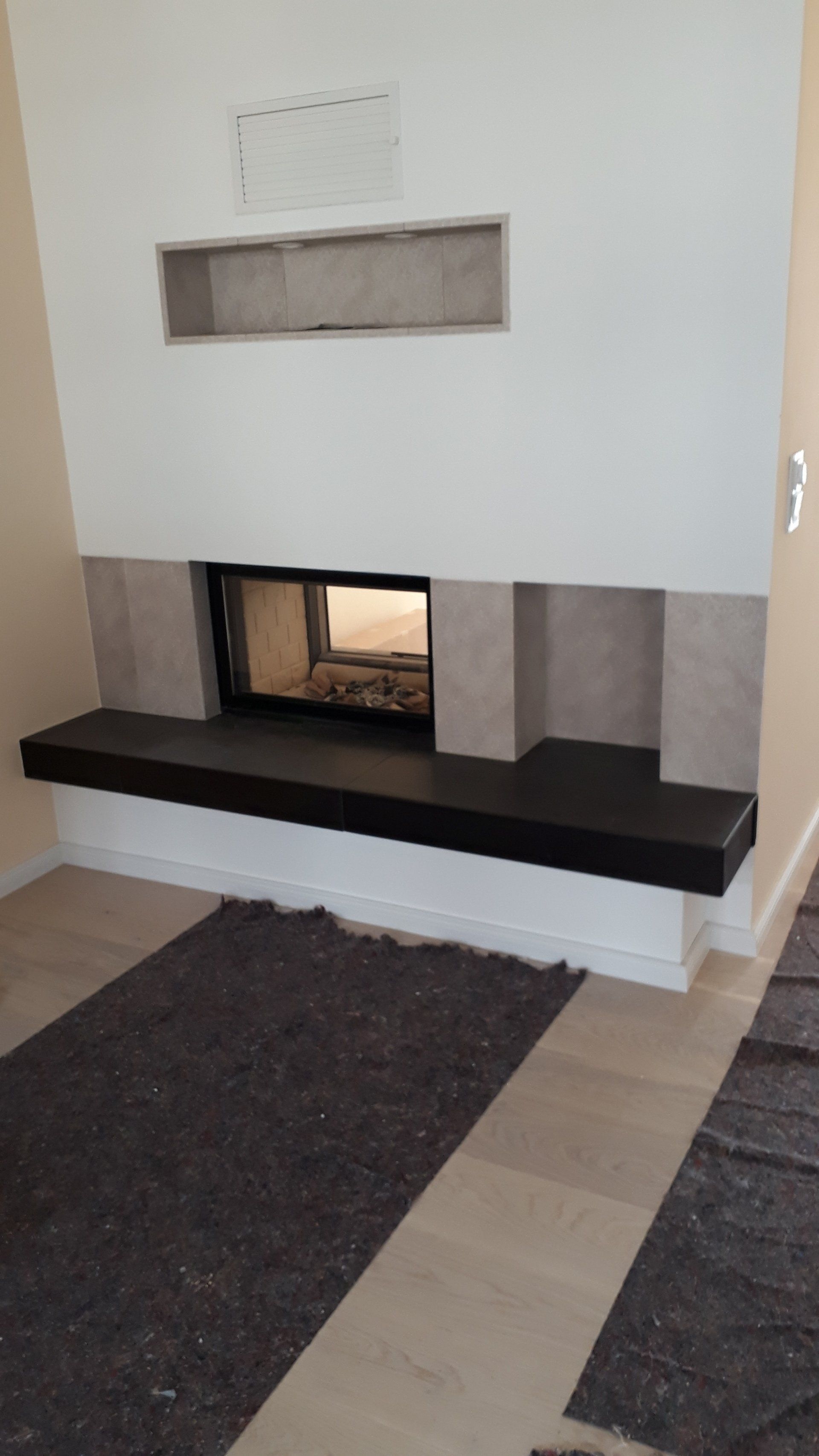 Kamin mit schwarzer Feuerstelle, eingebautem Feuerraum und einer Nische darüber, an einer weißen Wand.