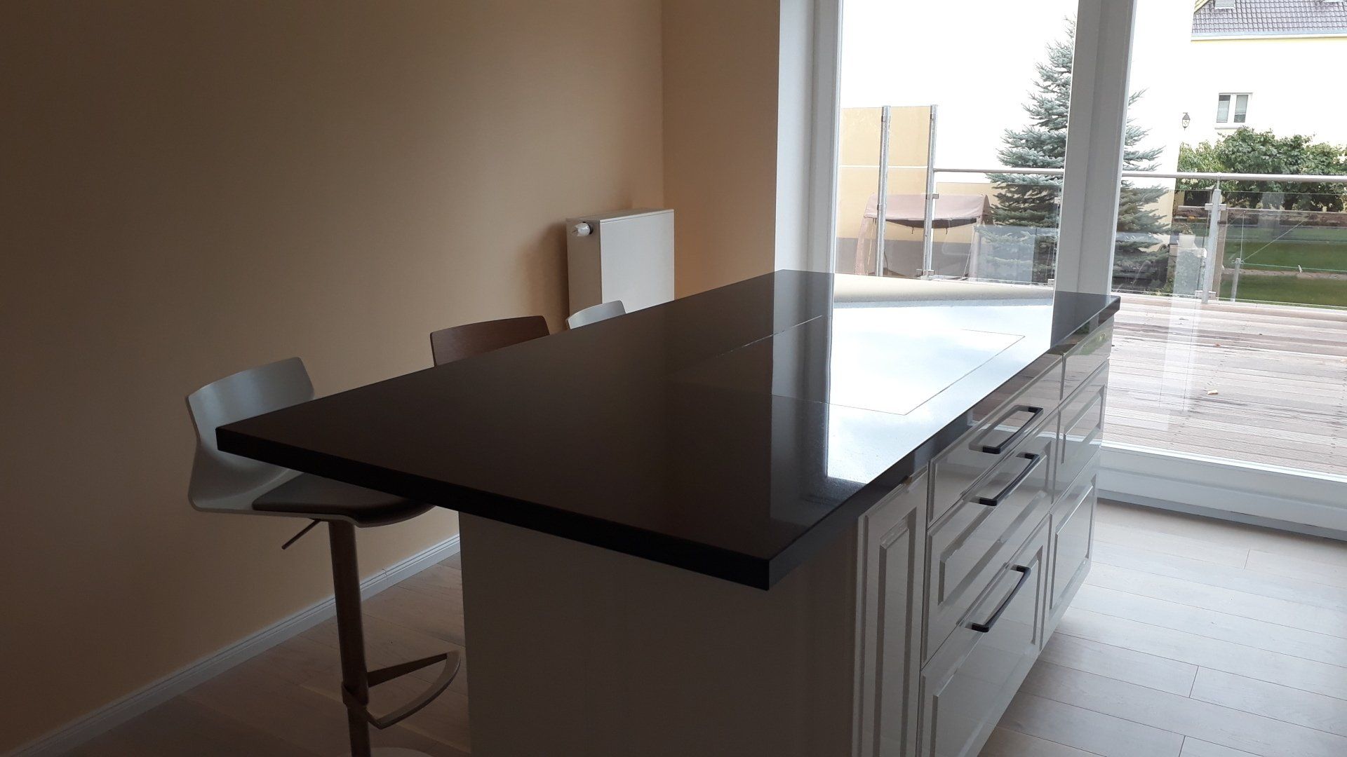 Kücheninsel mit dunkler Arbeitsplatte, weißen Schränken und zwei Barhockern. Neben einem Fenster.