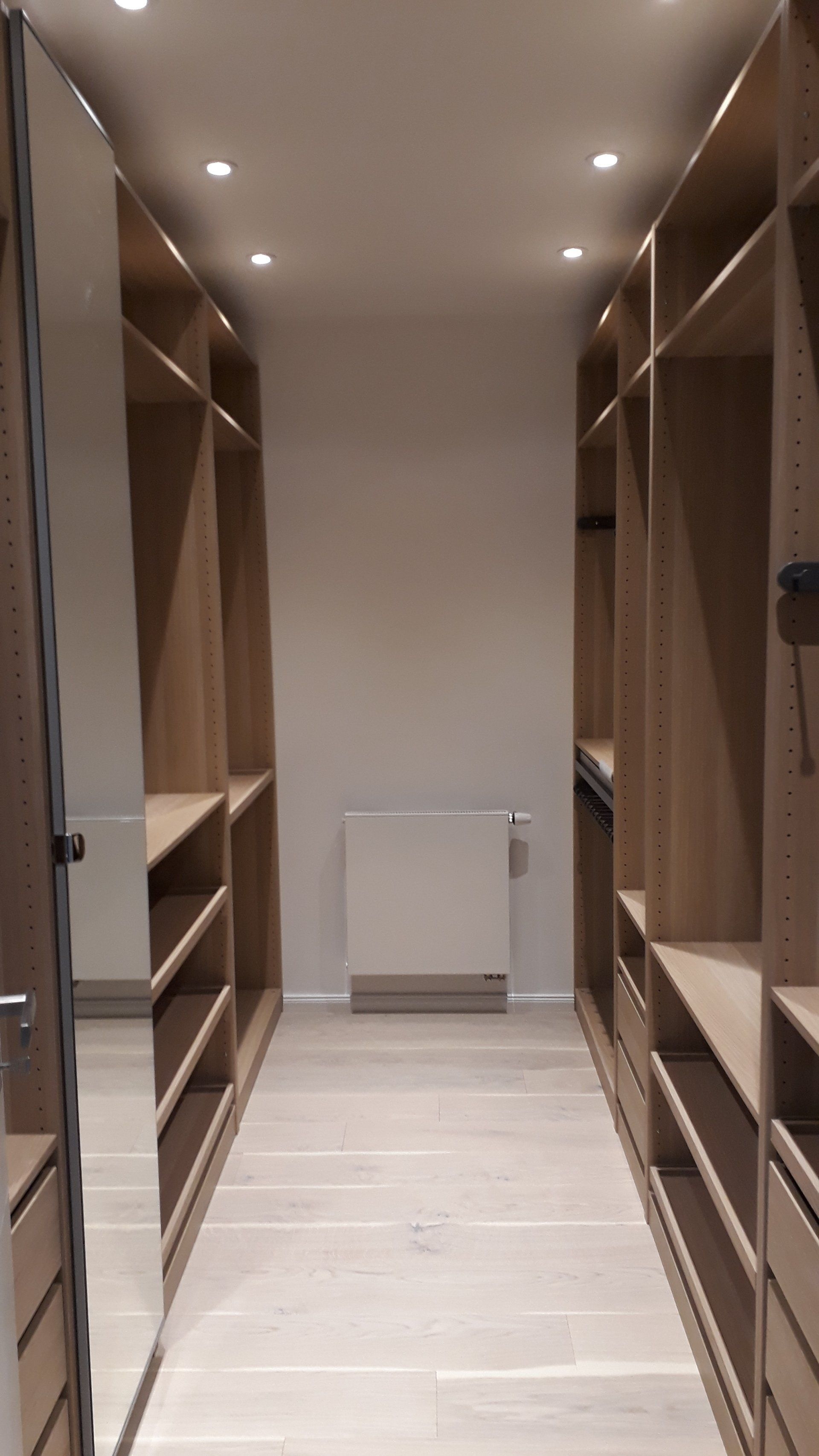 Begehbarer Kleiderschrank mit hellen Holzregalen und -boden, weißen Wänden und einem großen Spiegel.