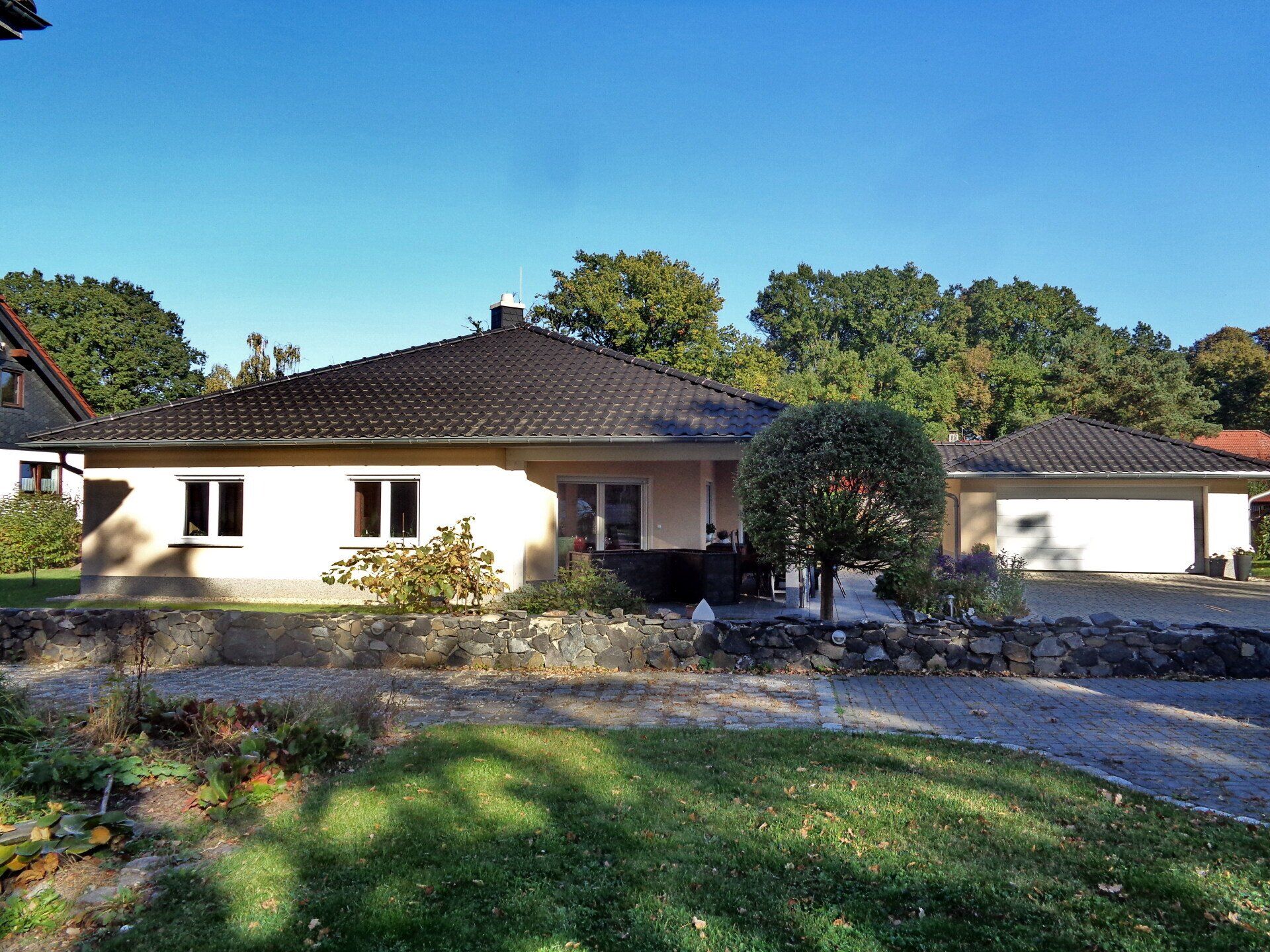 Beiges, eingeschossiges Haus mit schwarzem Dach und angebauter Garage, vor Bäumen, an einem sonnigen Tag.