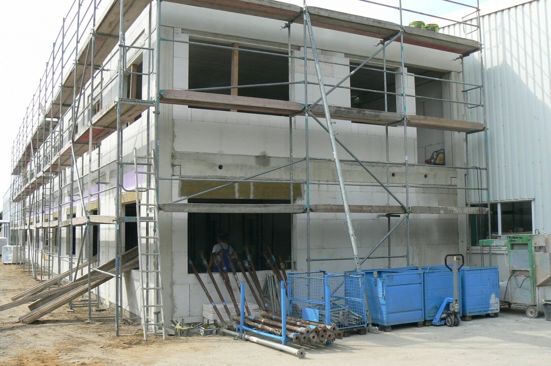 Ein im Bau befindliches Gebäude ist eingerüstet. Betonwände, Fensteröffnungen und blaue Mülltonnen.