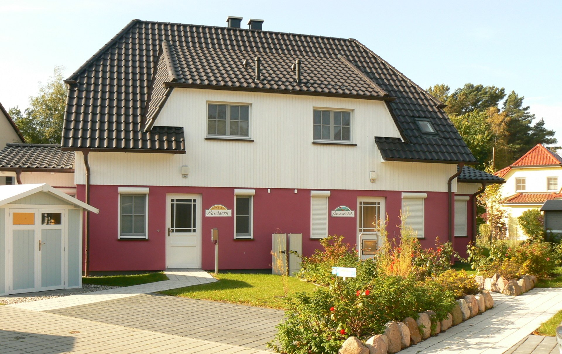 Zweistöckiges Haus mit schwarzem Dach und rosa-weißer Fassade. Gepflasterte Auffahrt, kleiner Garten mit Sträuchern.