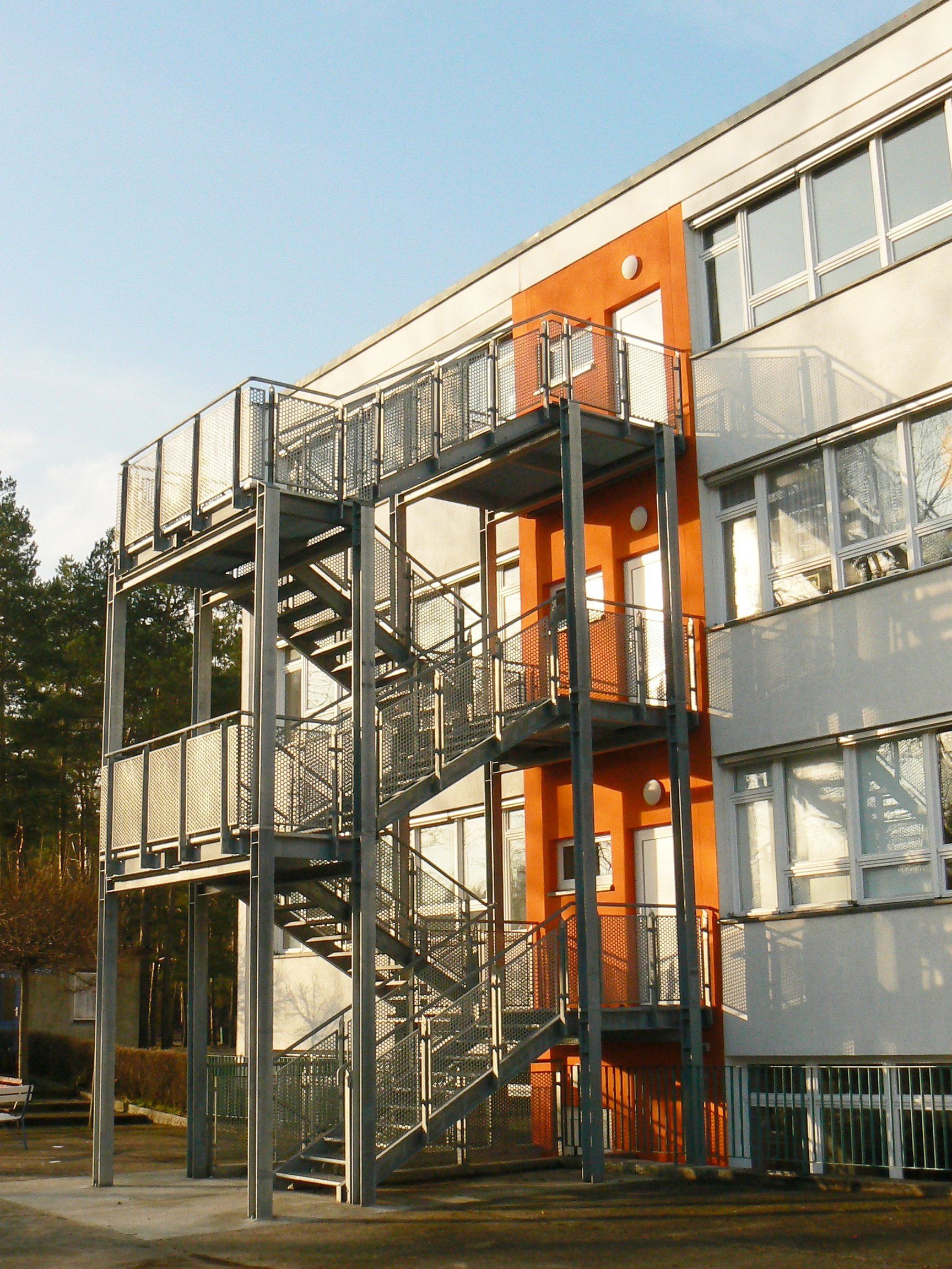 Außenansicht eines Gebäudes mit einer metallenen Feuertreppe. Oranger Wandabschnitt, Fenster und helles Sonnenlicht.