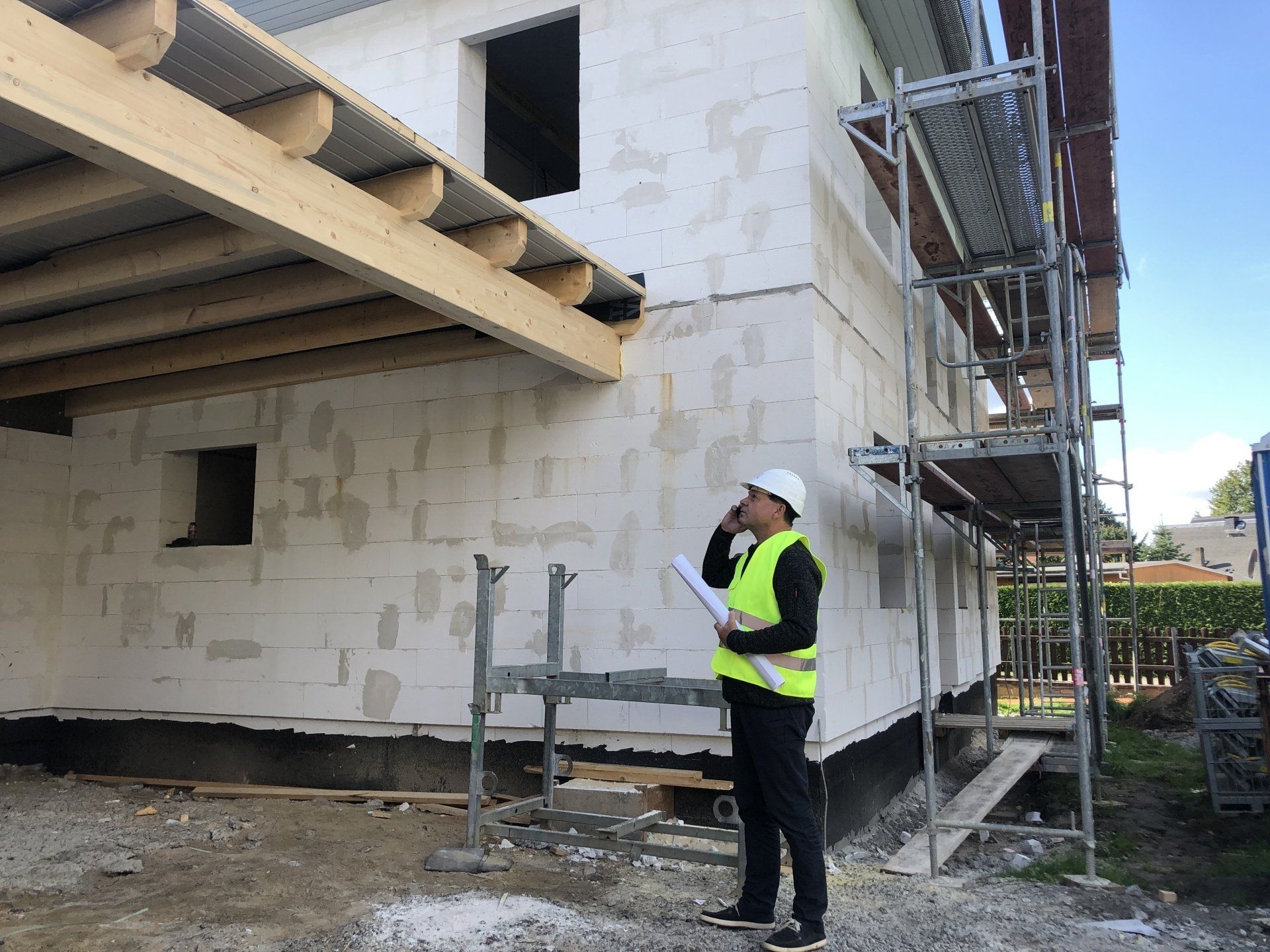 Ein Bauarbeiter mit Schutzhelm und Warnweste telefoniert und betrachtet ein im Bau befindliches Gebäude.