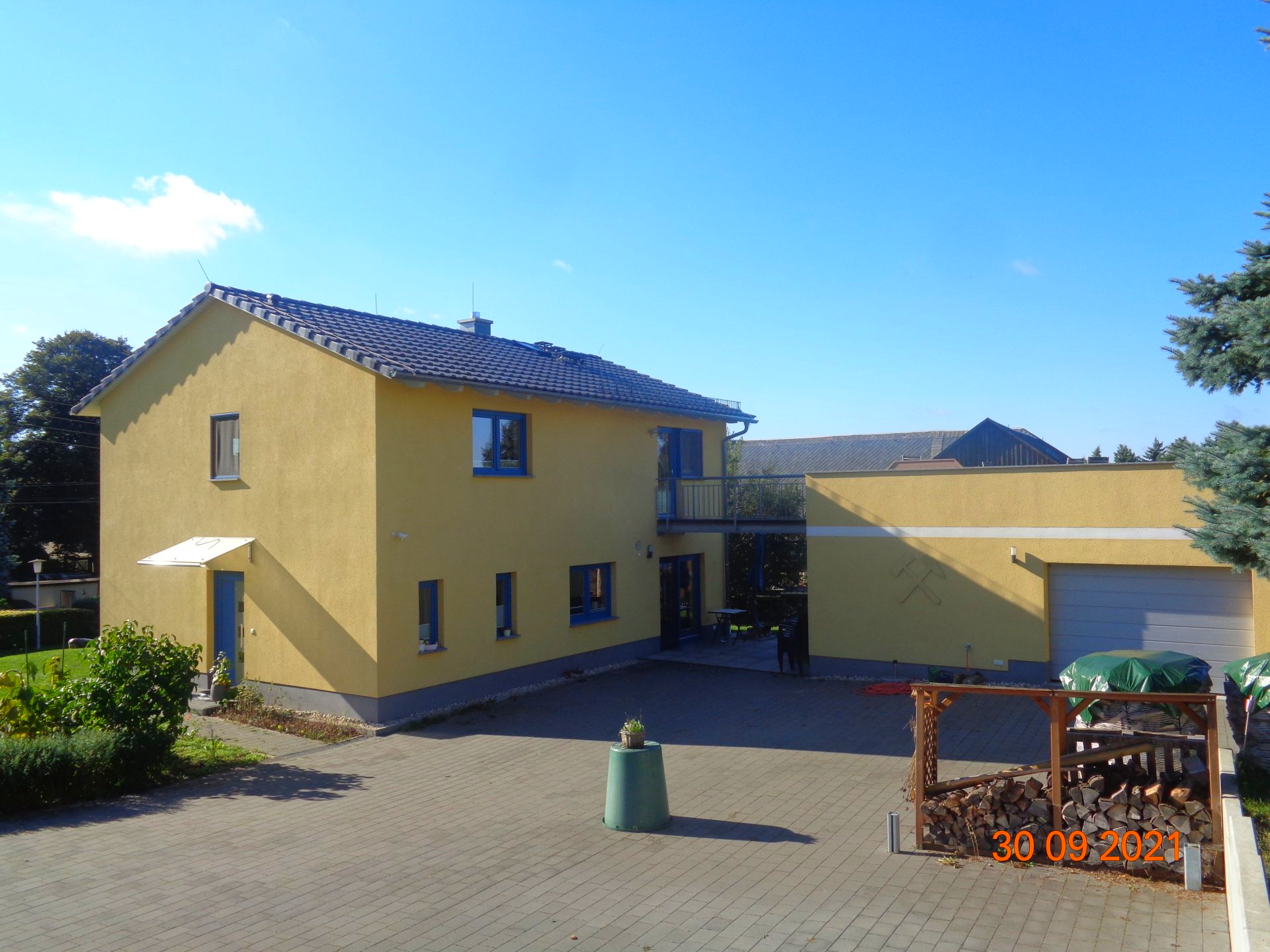 Gelbes zweistöckiges Haus mit Flachdach, Auffahrt und freistehender Garage vor blauem Himmel.