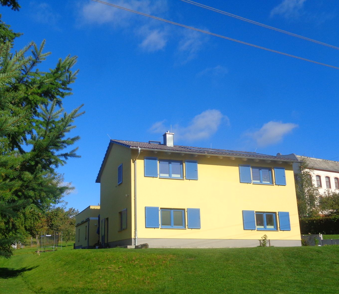 Gelbes Haus mit blauen Fensterläden auf einem grasbewachsenen Hügel unter strahlend blauem Himmel.