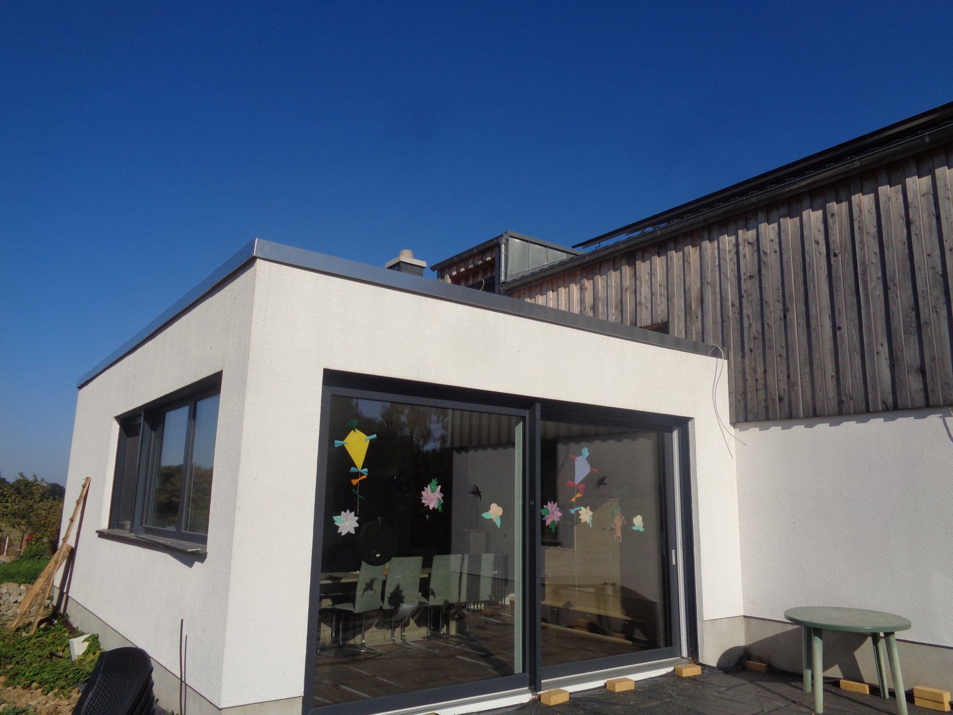 Weißes Gebäude mit großen Glastüren und -fenstern, neben einem Holzbau, vor blauem Himmel.