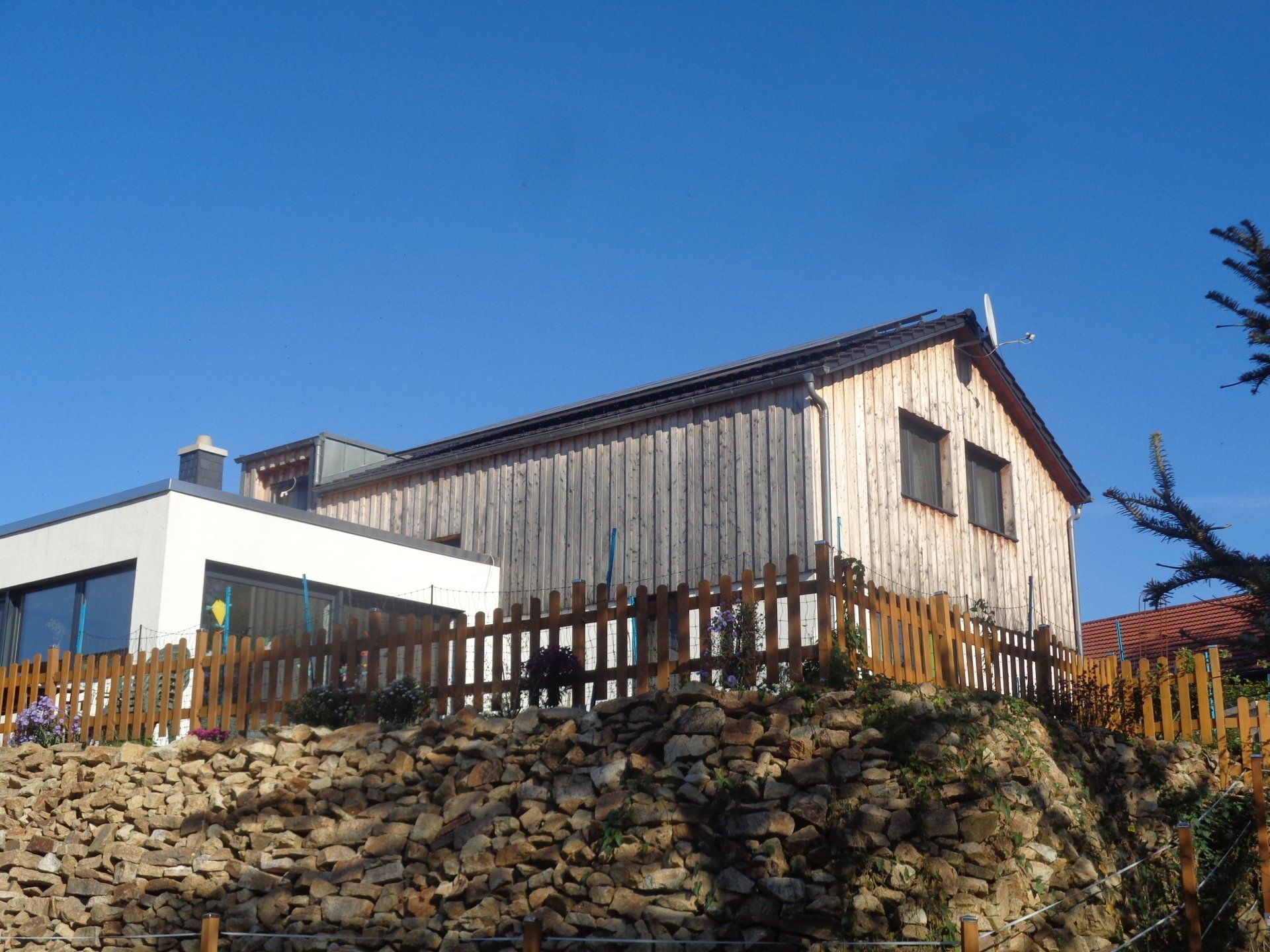 Zweistöckiges Holzgebäude auf einem Hügel mit einem kleinen Zaun, ein weißes Nebengebäude, unter blauem Himmel.