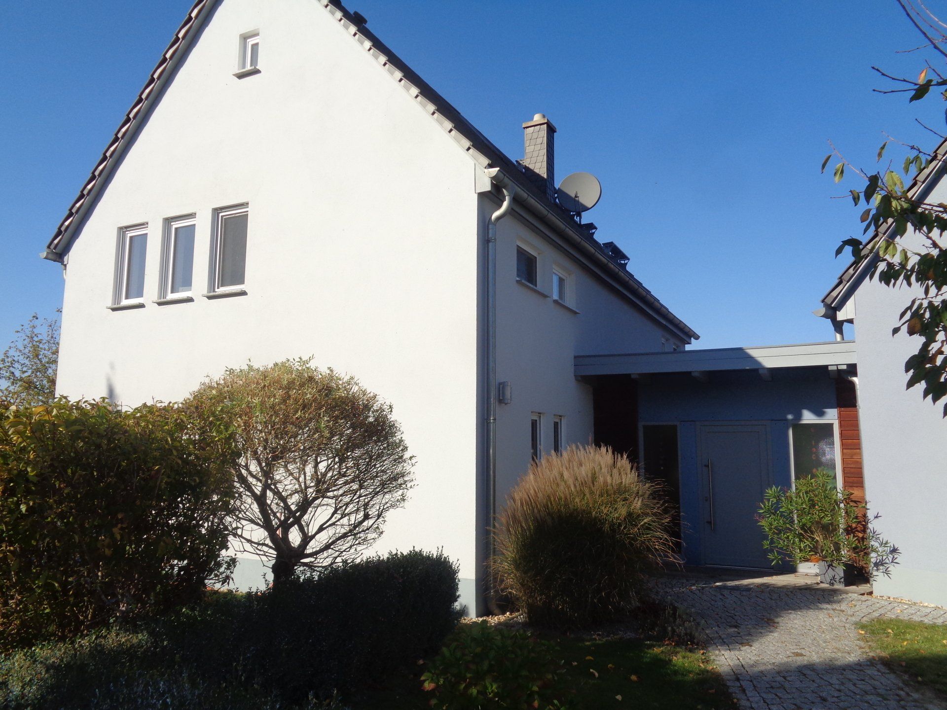 Weißes Haus mit grauem Anbau, Büschen davor und blauem Himmel.