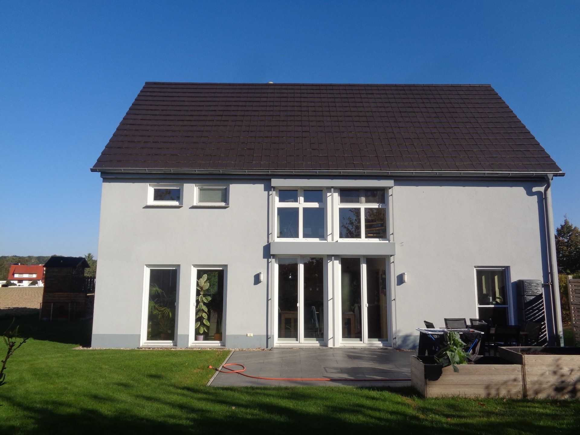 Graues zweistöckiges Haus mit dunklem Dach vor klarem blauem Himmel, auf einem grünen Rasen gelegen.