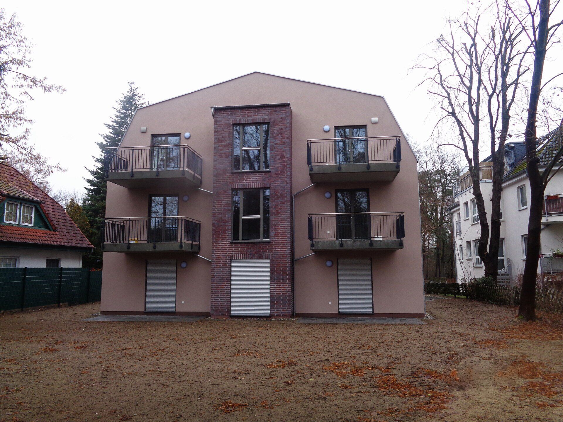 Mehrfamilienhaus mit Balkonen und einem zentralen Backsteinbau. Beige Fassade, braune Türen, kahle Bäume.