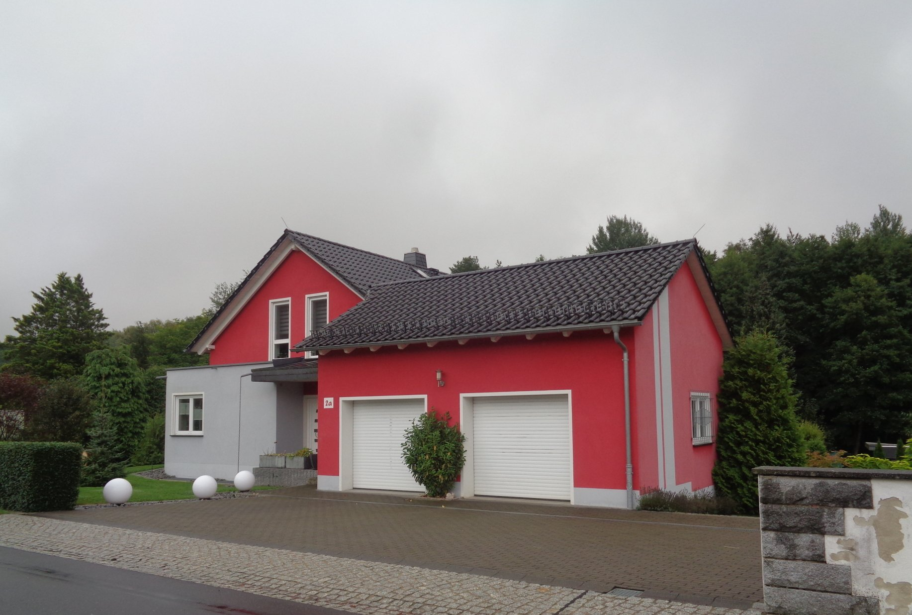 Rotes Haus mit Garage, weißen Türen und dunklem Dach unter bewölktem Himmel.