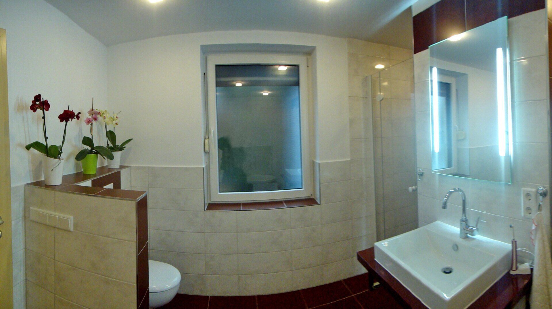 Modernes Badezimmer mit Fenster, Orchideen, quadratischem Waschbecken und roten und beigen Fliesen.