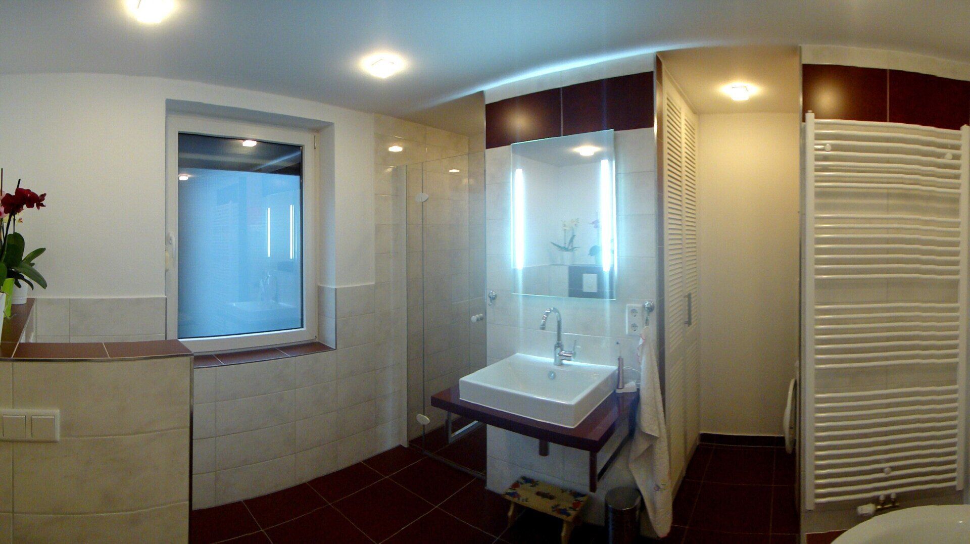 Badezimmer mit weißem Waschbecken, Dusche und Handtuchwärmer. Rotbrauner Boden und Akzentfarben.