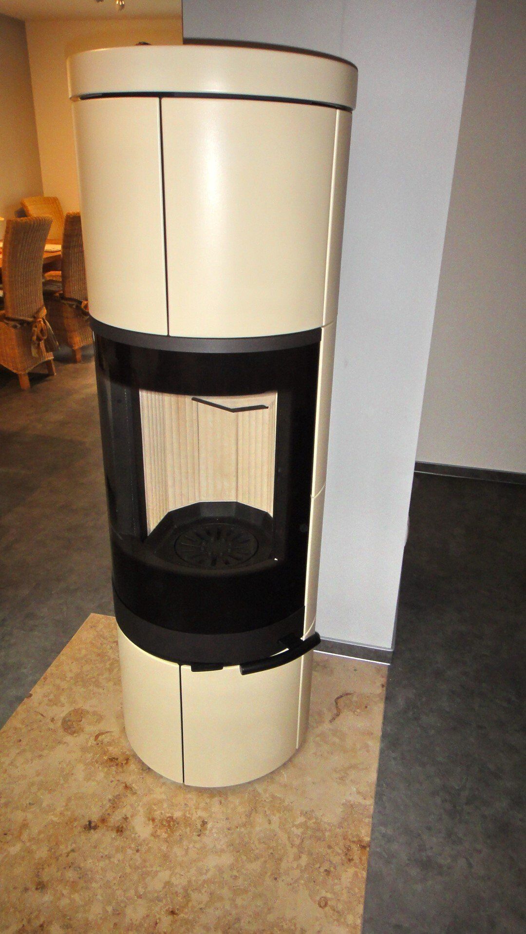 Ein cremefarbener, zylindrischer Kamin mit einem schwarz umrandeten Sichtfenster. Er steht auf einem braunen Steinsockel und befindet sich im Innenbereich.