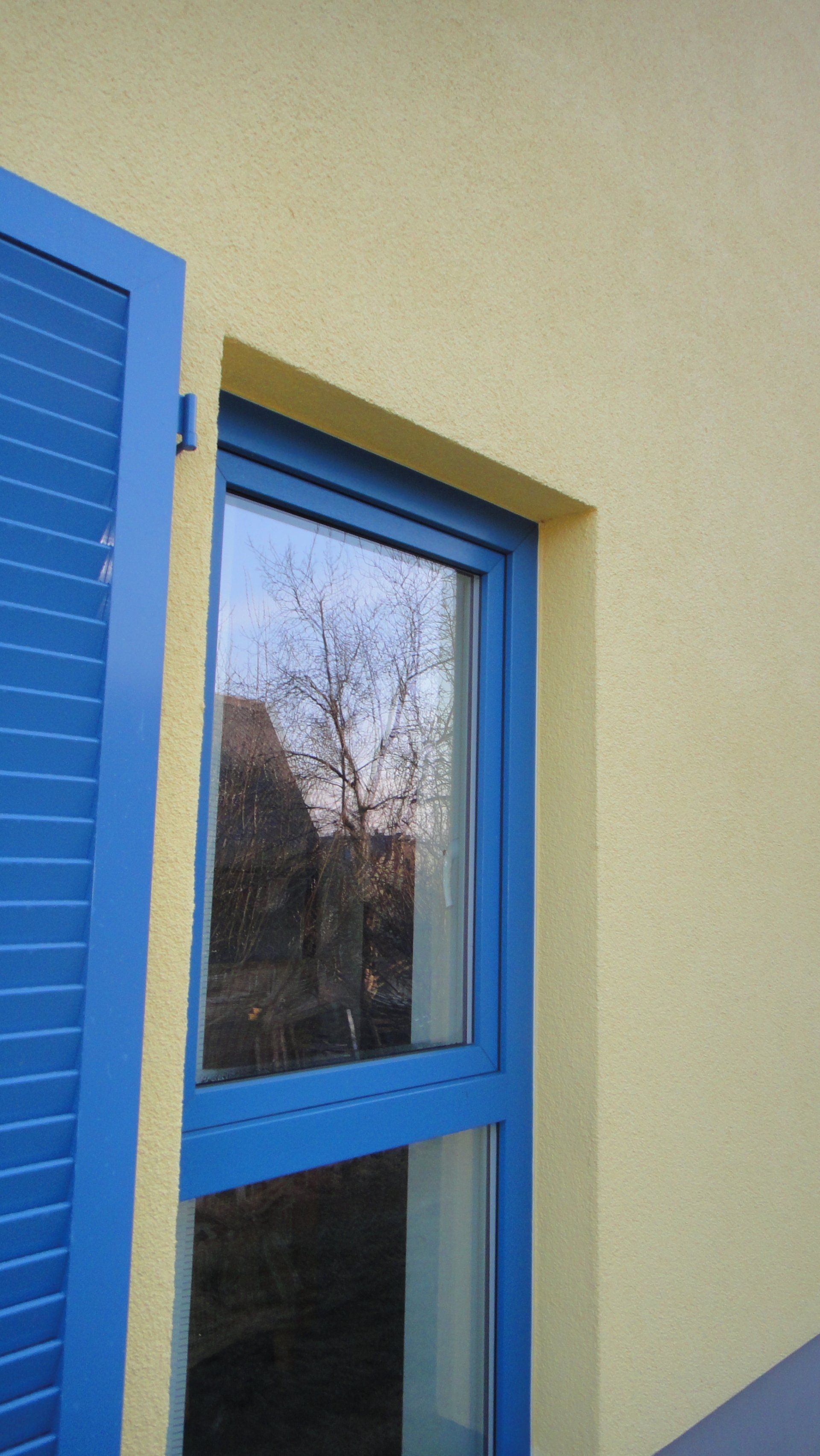 Blau umrahmtes Fenster mit blauem Fensterladen an einer gelben Stuckwand, in der sich Bäume spiegeln.